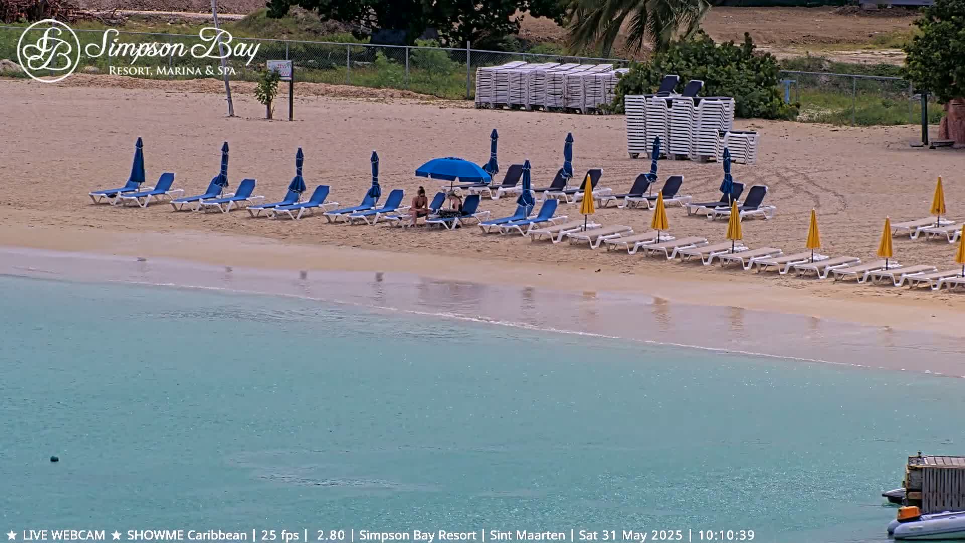 A sandy beach with numerous lounge chairs and umbrellas, two people sitting under one umbrella, and calm, clear turquoise water.