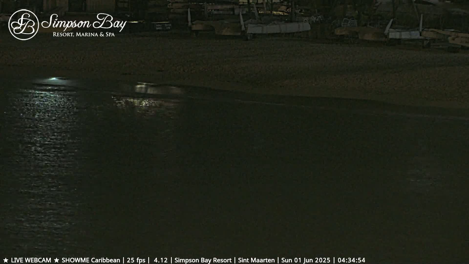 At night, several boats are docked on a dark, sandy shore next to calm, dark water reflecting light.