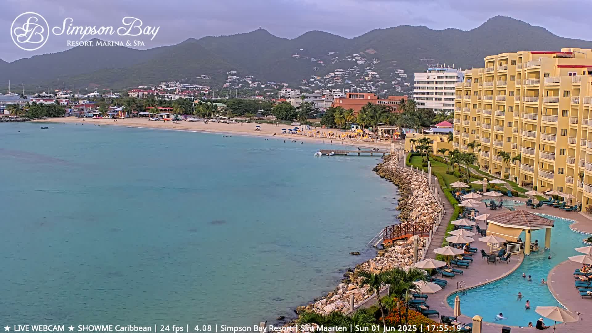 A beach with turquoise water, a sandy shoreline, people swimming and lounging, a resort with a pool and multiple buildings, and mountains in the background under an overcast sky.