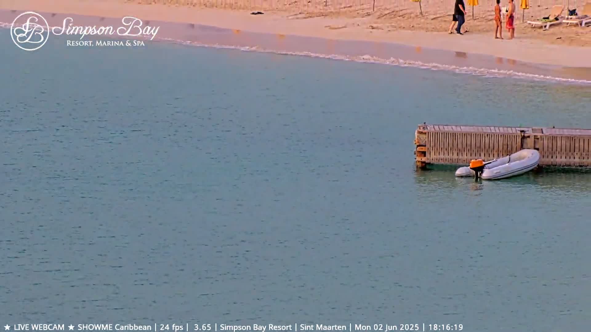 A small inflatable motorboat is moored next to a wooden dock in calm, light-blue water near a sandy beach with a few people.