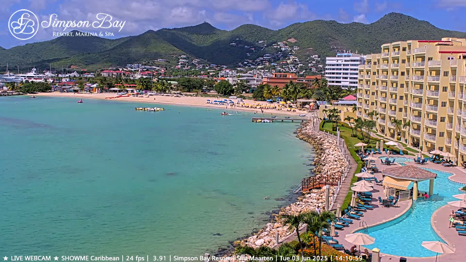 A tropical beach scene with turquoise water, a sandy shore with beach umbrellas, a large resort building with a pool, and lush green hills in the background under a sunny sky.
