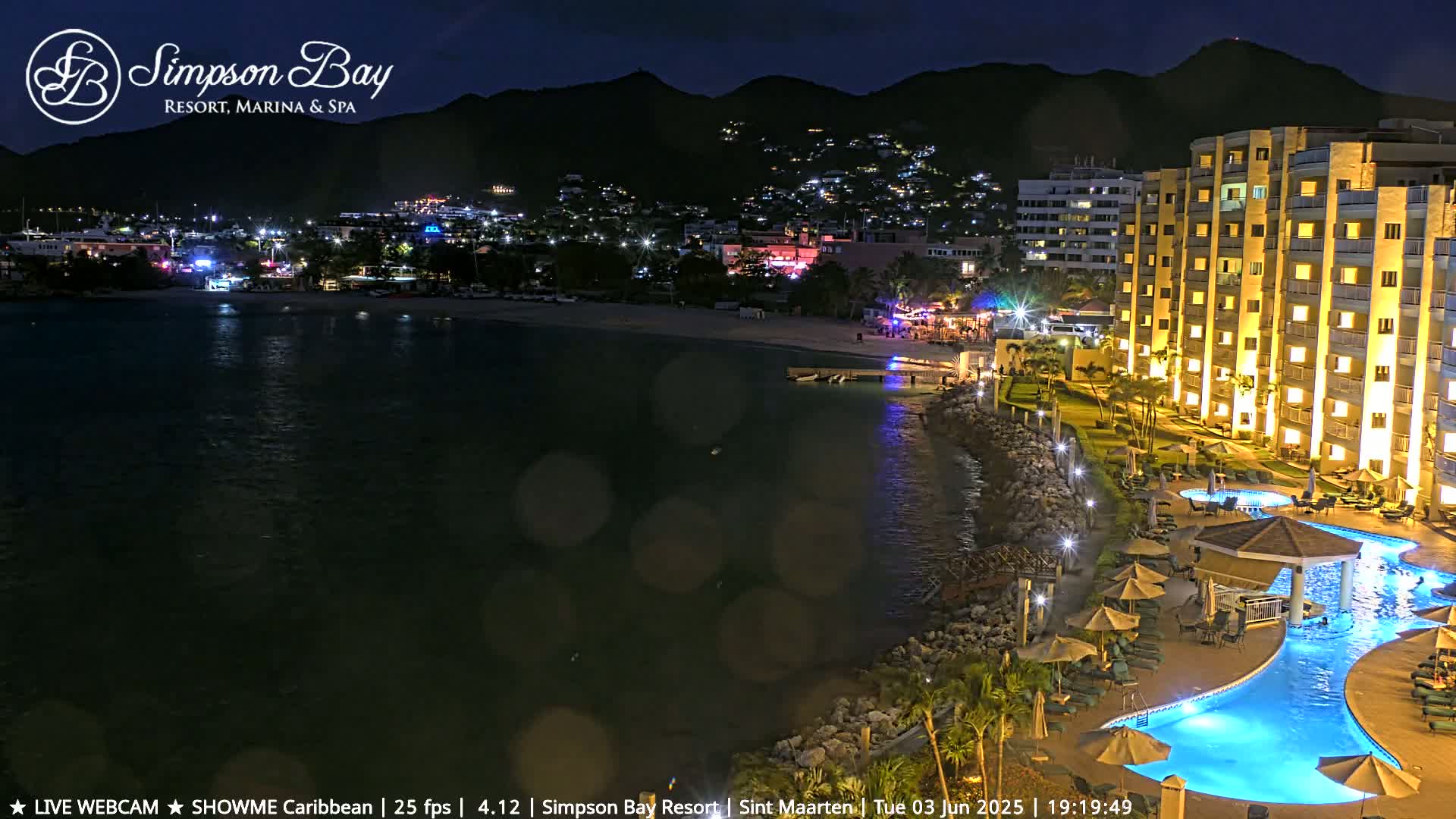 A brightly lit resort with a large pool sits on a coast at night, overlooking a dark body of water and a town illuminated with lights against a mountainous backdrop.