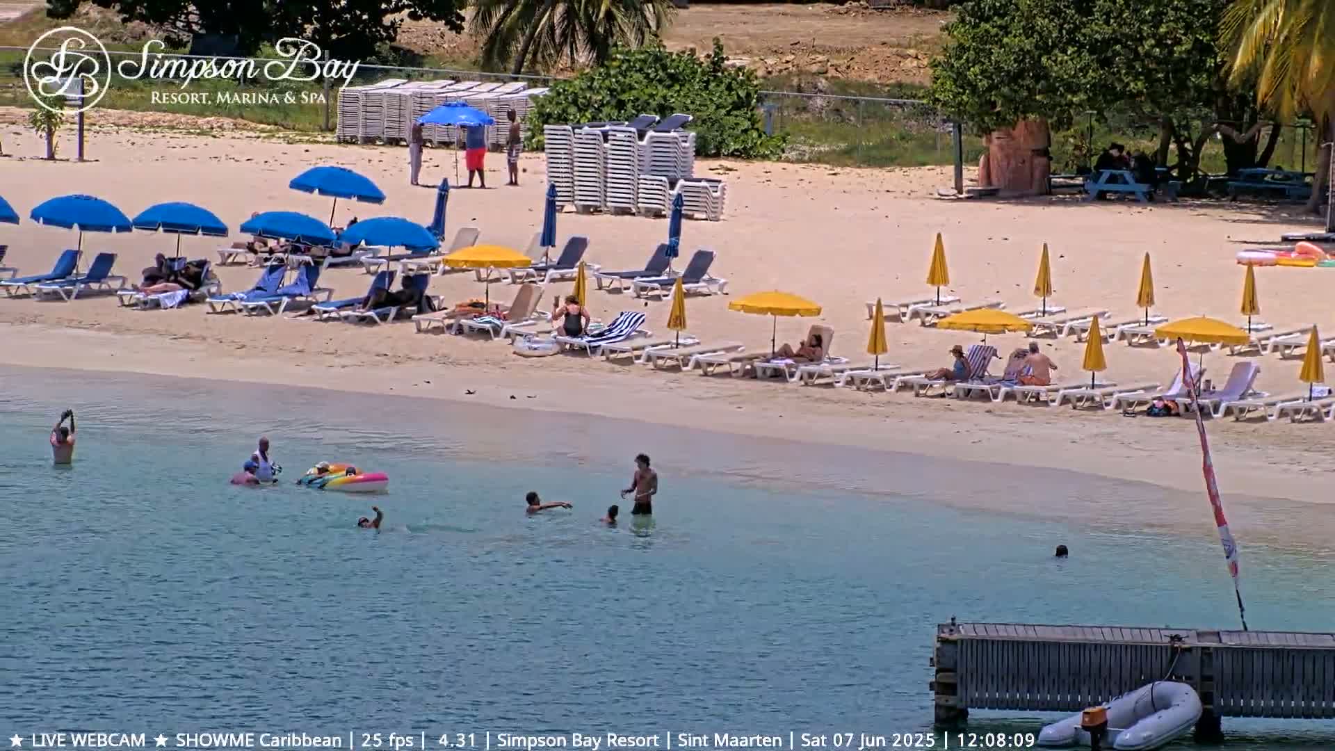 A sunny beach scene shows people relaxing on lounge chairs under umbrellas, with others swimming in calm, shallow water near a small dock.