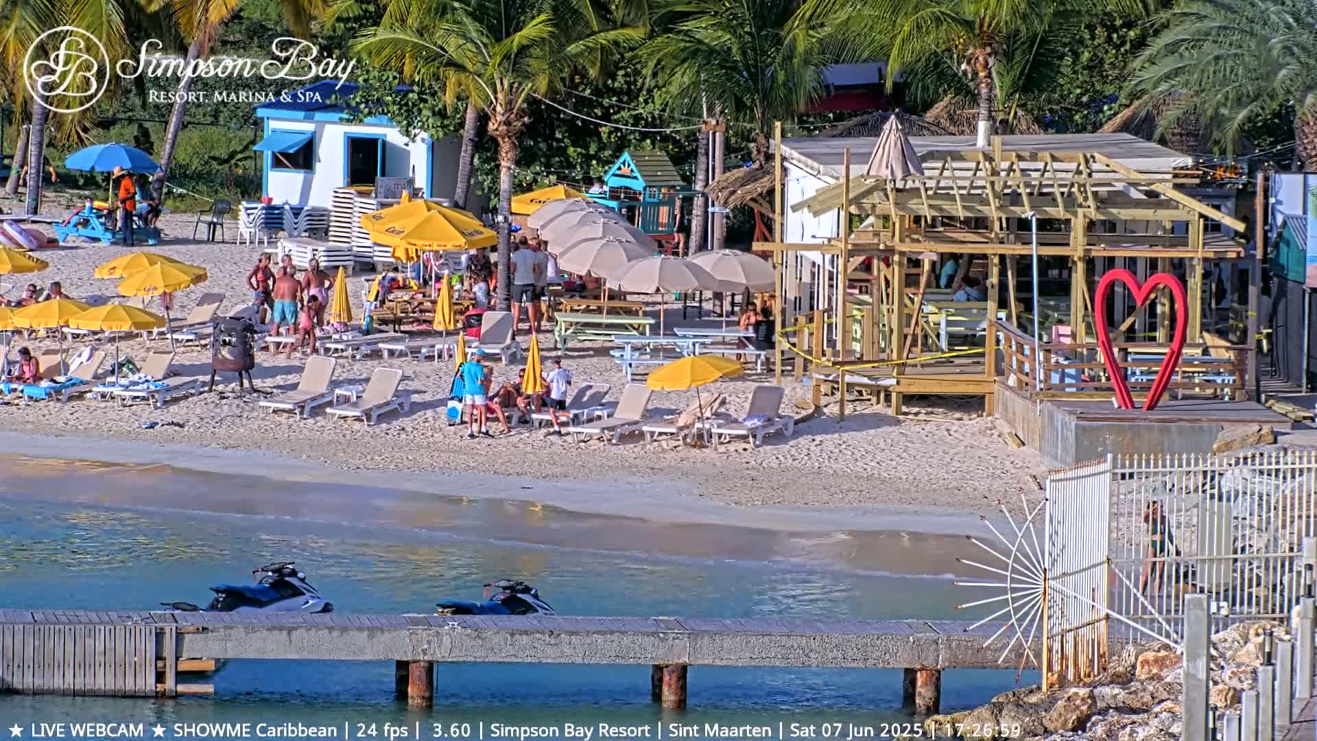 A beach scene on a sunny day shows people relaxing under umbrellas near a beachfront restaurant under construction, with jet skis docked nearby.