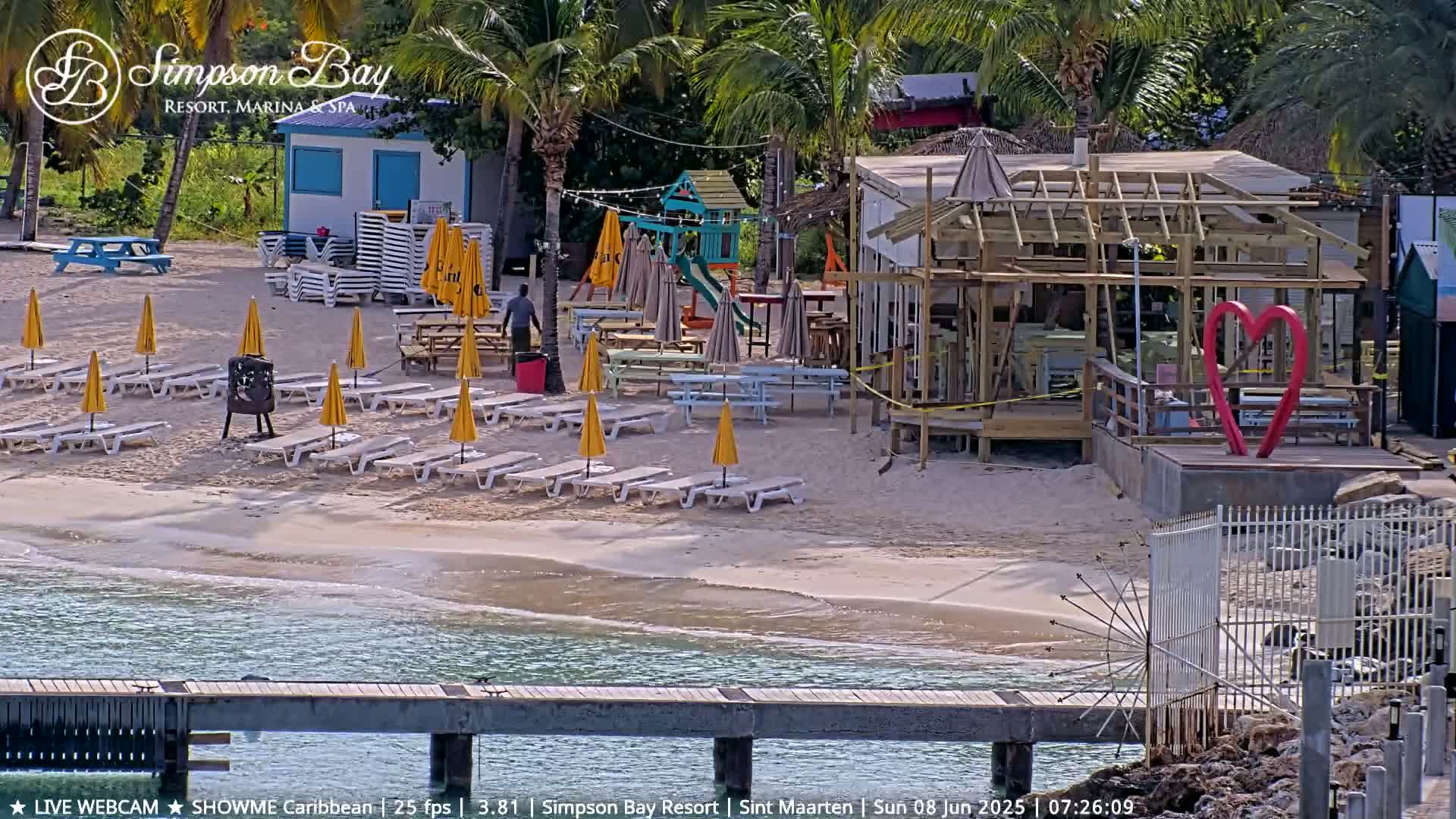 A sandy beach with numerous yellow umbrellas and lounge chairs, a playground, a partially constructed building, and a large red heart sculpture sits near calm, shallow blue water.