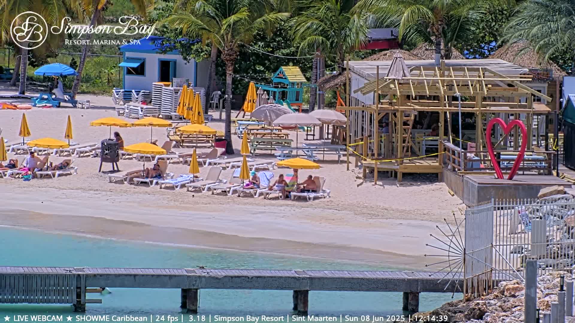 A sunny beach scene shows people relaxing on lounge chairs under yellow umbrellas near a partially constructed building with a large red heart sculpture, next to calm, turquoise water and a wooden pier.