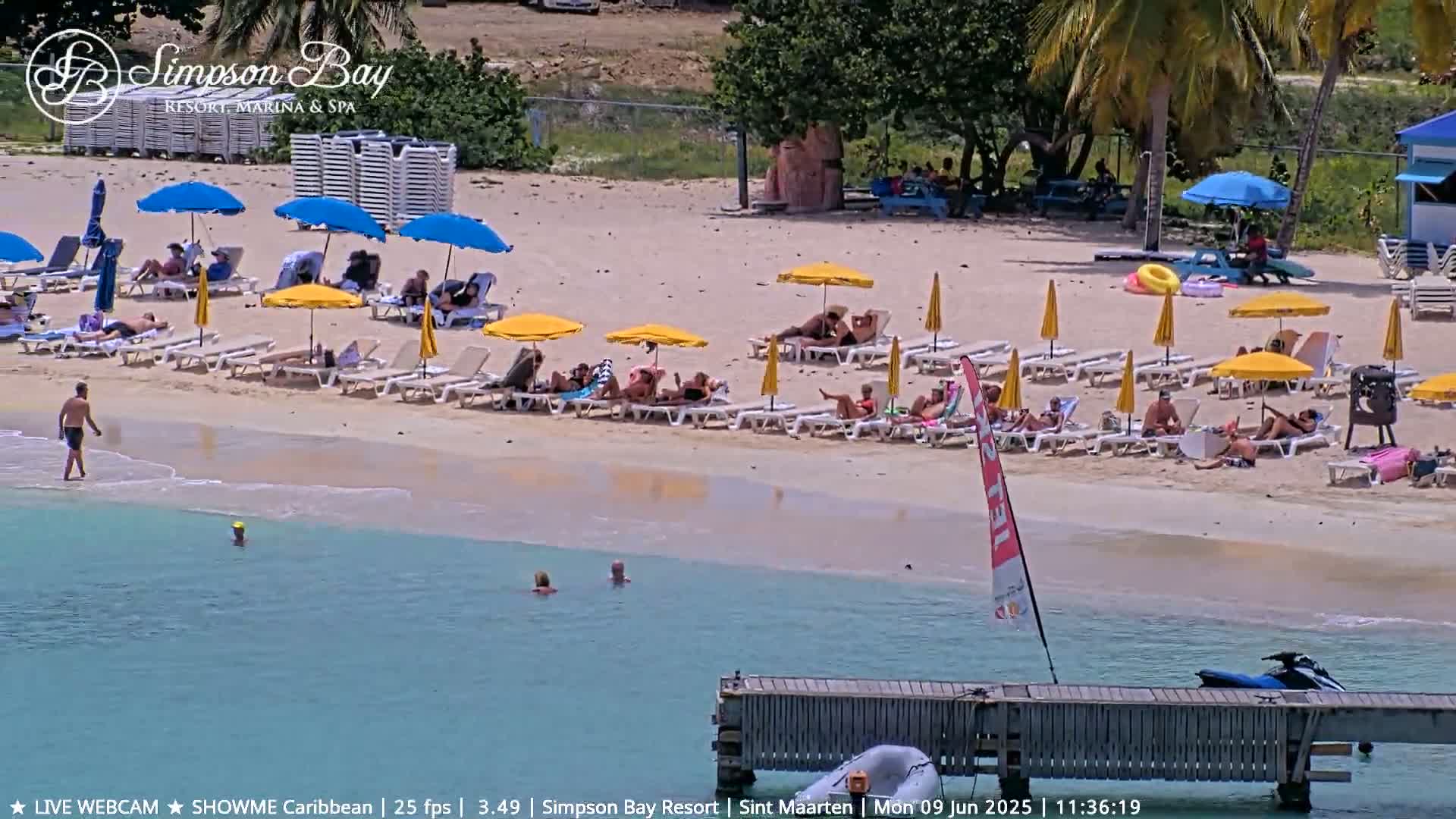A sunny beach scene shows people relaxing on lounge chairs under umbrellas, with several individuals swimming in calm, turquoise water near a pier and a jet ski.