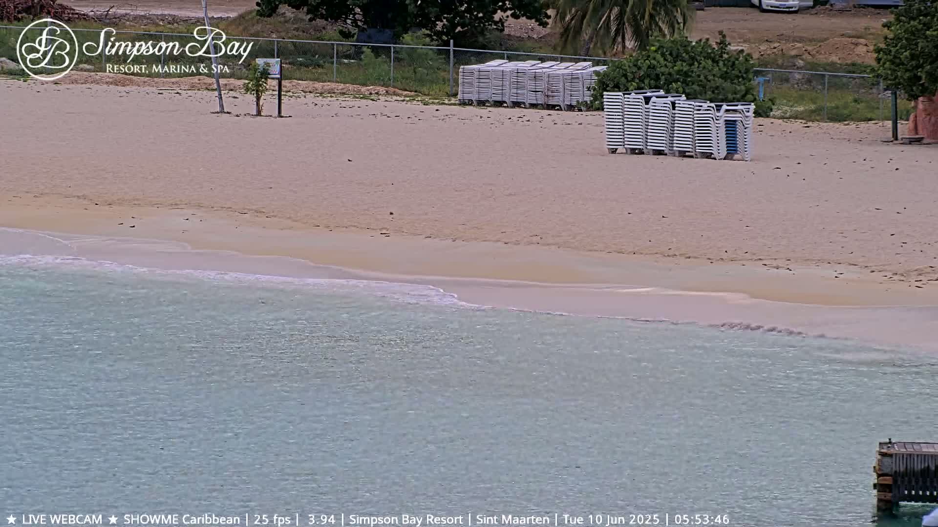 A sandy beach with calm, light-blue water laps gently at the shore, and stacks of white lounge chairs sit on the sand.