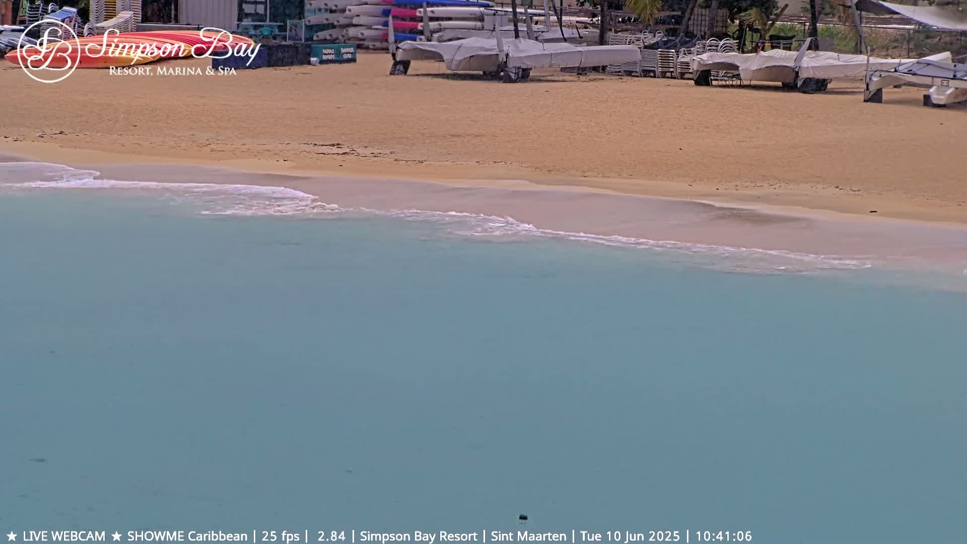 A sandy beach with calm, light-blue water laps gently at the shore, with several boats and kayaks visible on the beach in the background.