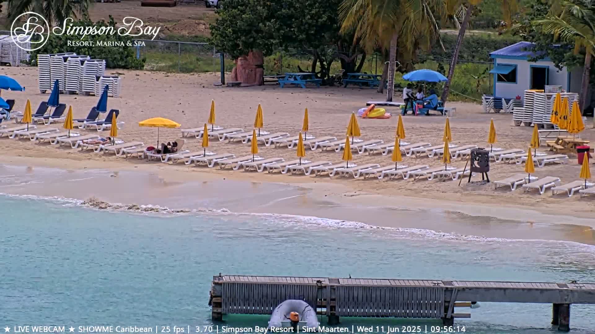 A sandy beach with numerous yellow umbrellas and lounge chairs next to calm, turquoise water, a small dock, and some palm trees.