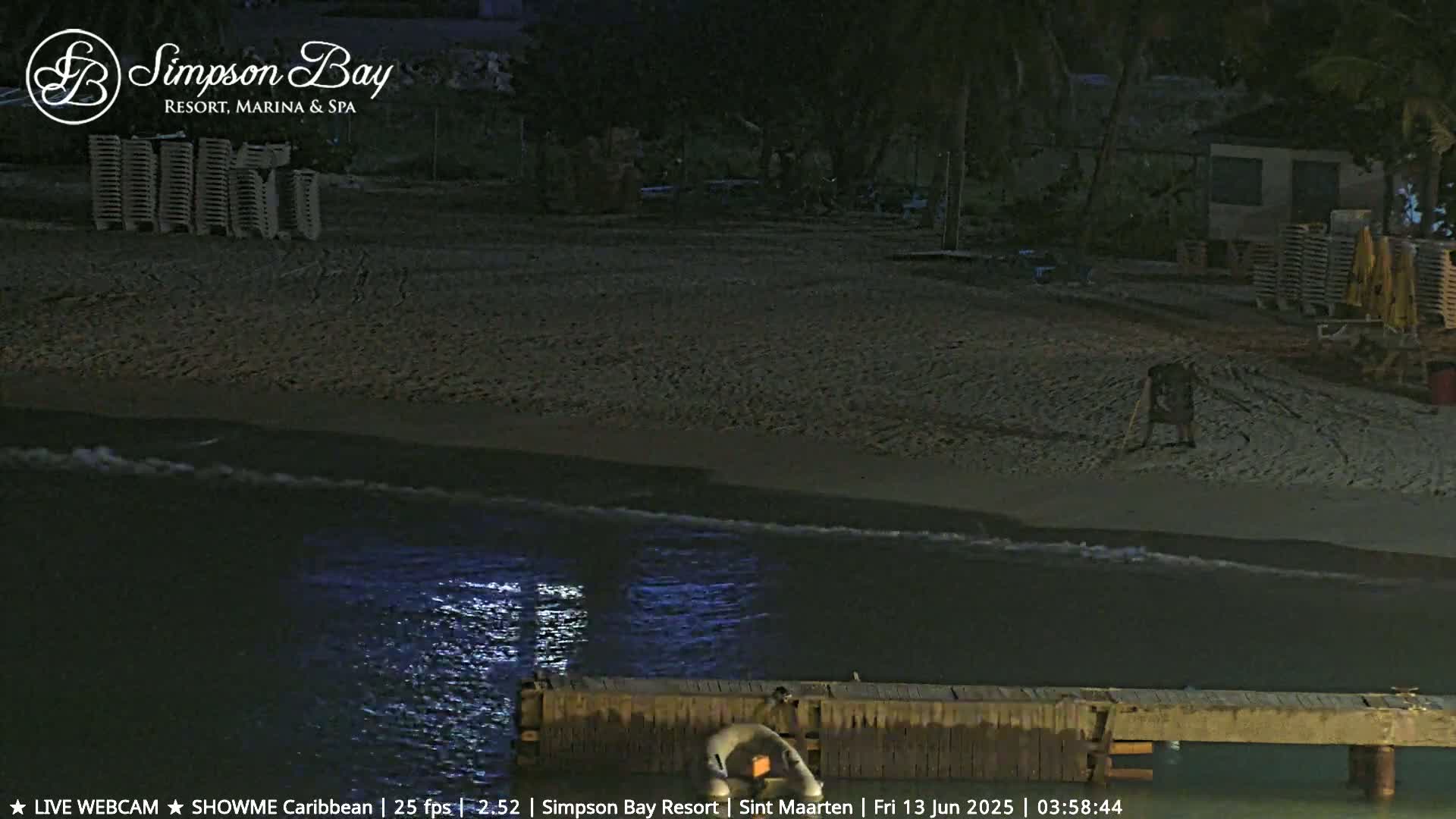 A nighttime beach scene shows a dark sandy shore with calm waves lapping at a wooden pier; several lounge chairs are stacked in the background.