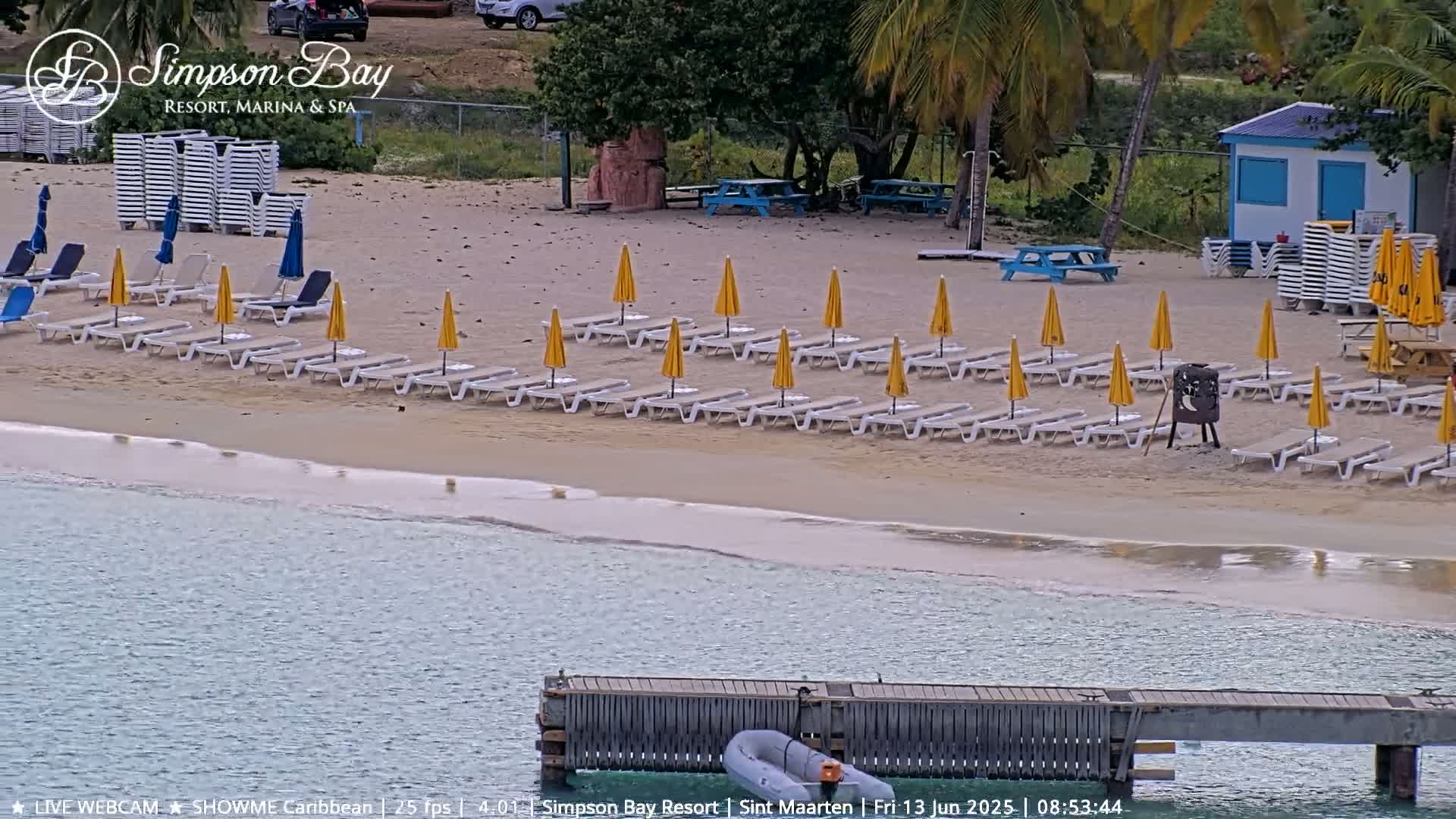 A sandy beach with numerous lounge chairs and yellow umbrellas under a partly sunny sky, next to calm, clear water and a small dock with a dinghy.
