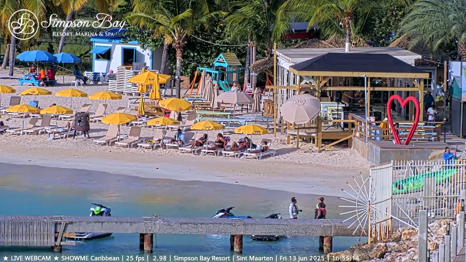 A sunny beach scene shows numerous yellow umbrellas and beach chairs surrounding a beach bar, with people relaxing and jet skis near a pier in calm turquoise water.