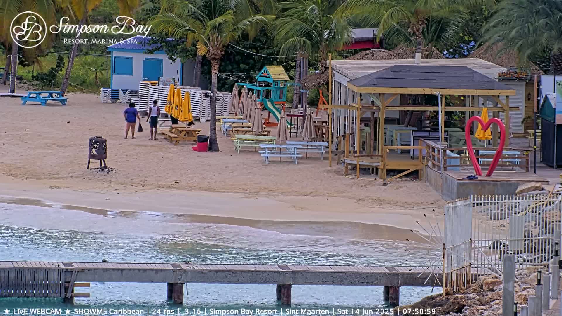 A sandy beach with picnic tables, a playground, and a beachfront bar and restaurant is shown, with calm water lapping the shore.