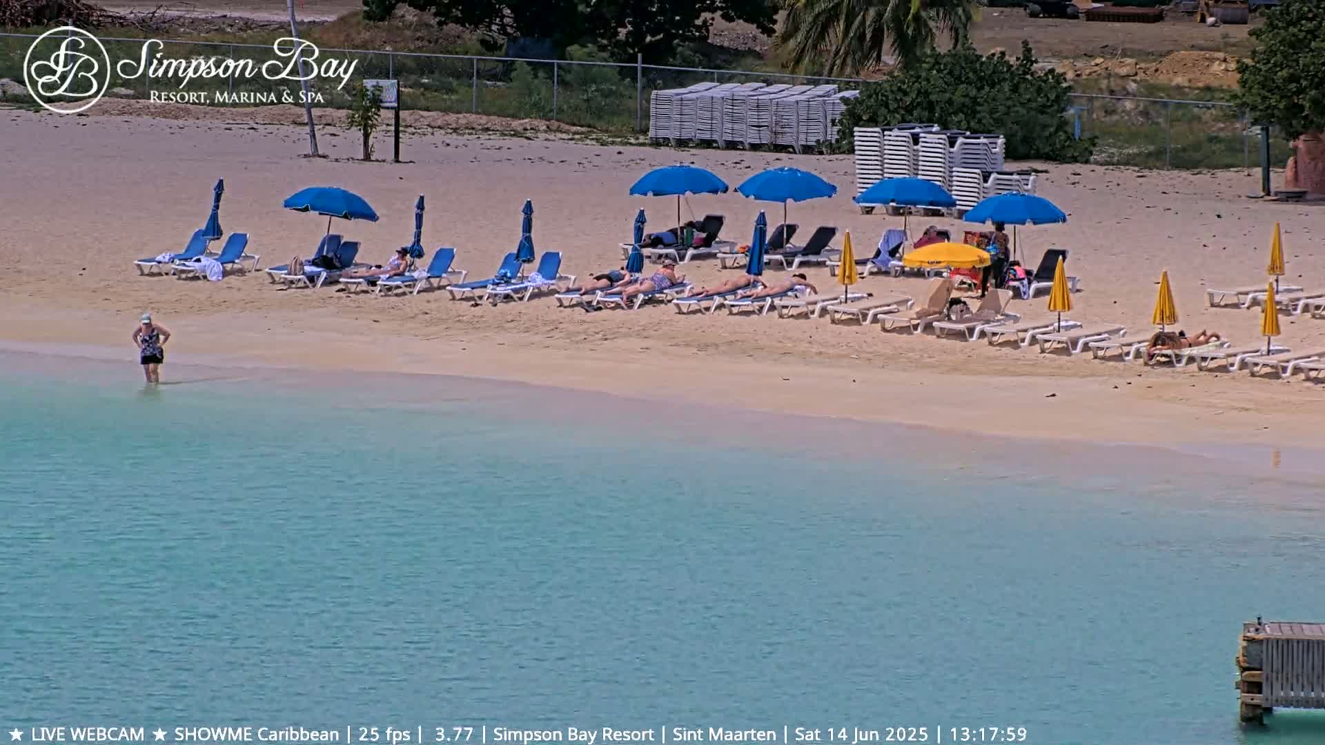 A sandy beach with turquoise water features numerous beach chairs and umbrellas under a sunny sky, with several people sunbathing and one person wading in the shallows.