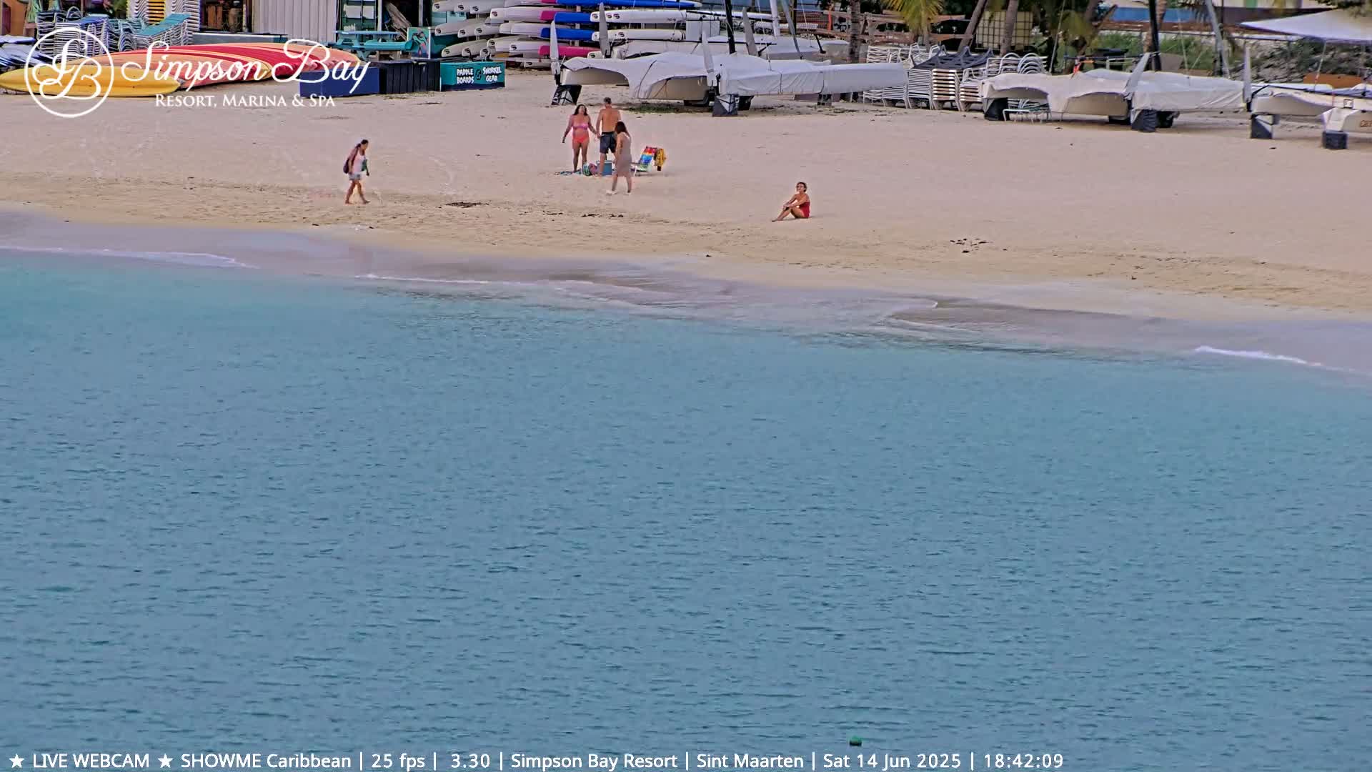 A sandy beach with turquoise water features numerous beach chairs and umbrellas under a sunny sky, with several people sunbathing and one person wading in the shallows.
