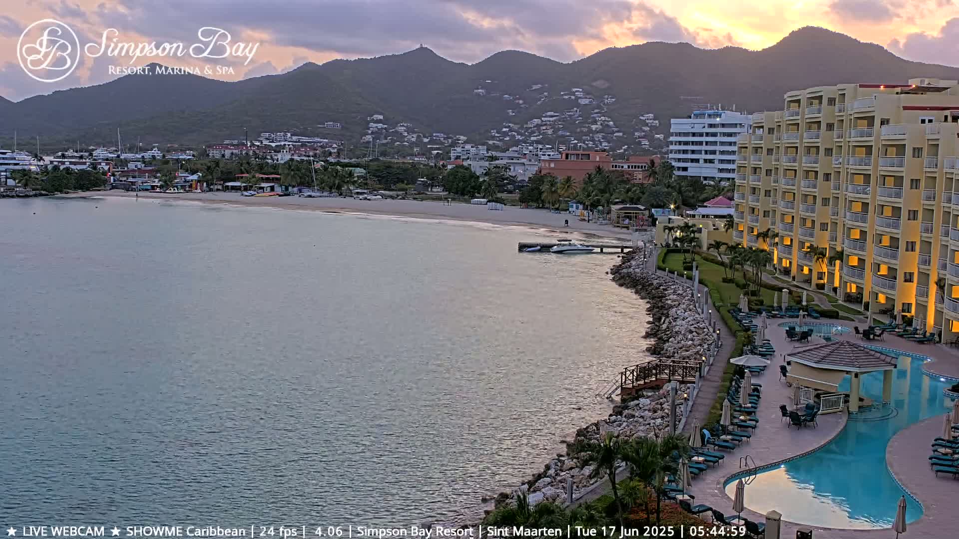 A calm bay with a sandy beach, resort buildings, and a swimming pool is seen at sunset, with hills in the background.
