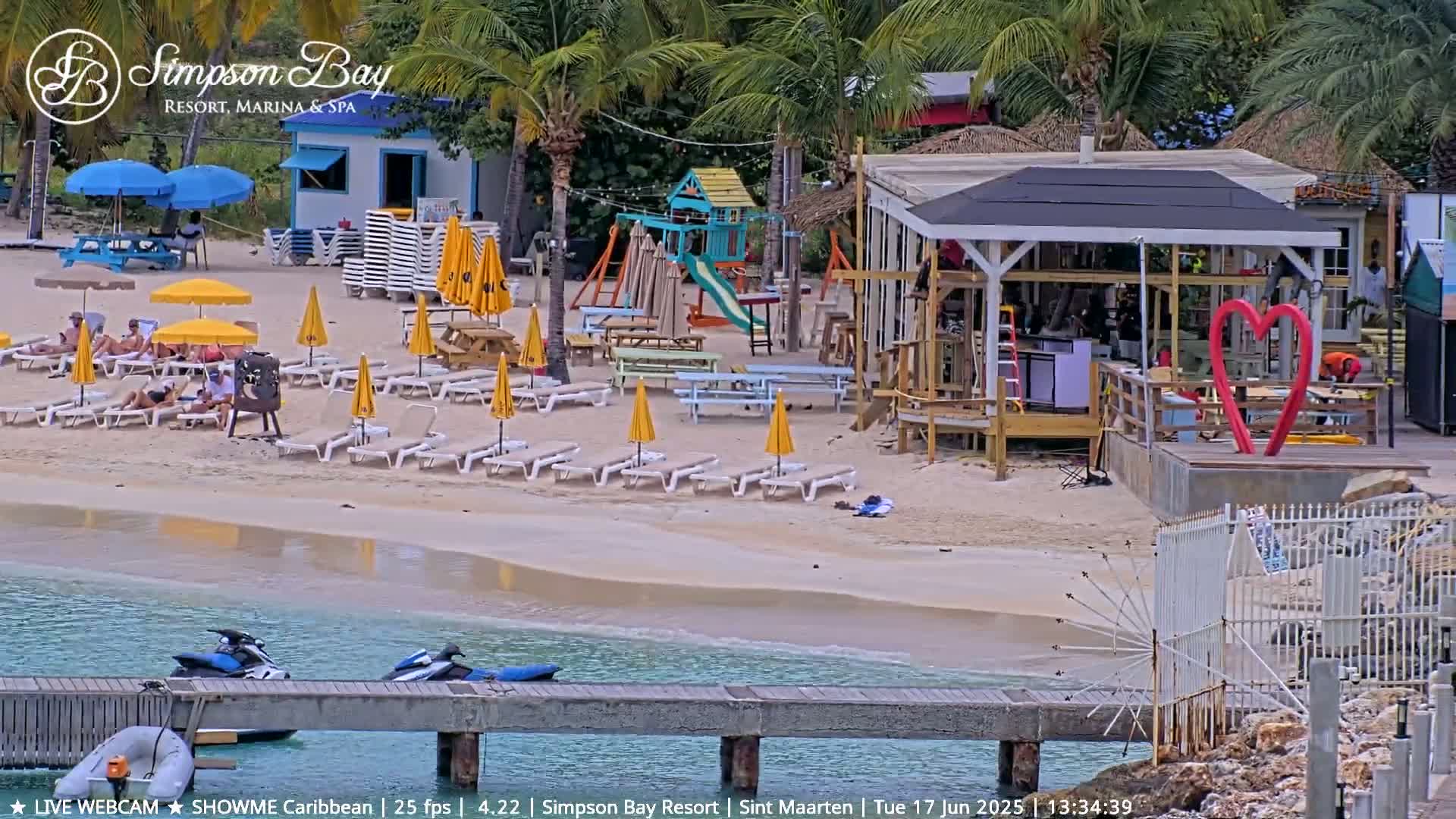 A beach scene shows lounge chairs under yellow umbrellas, a beach bar, a playground, and jet skis at a dock, on a calm, sunny day.