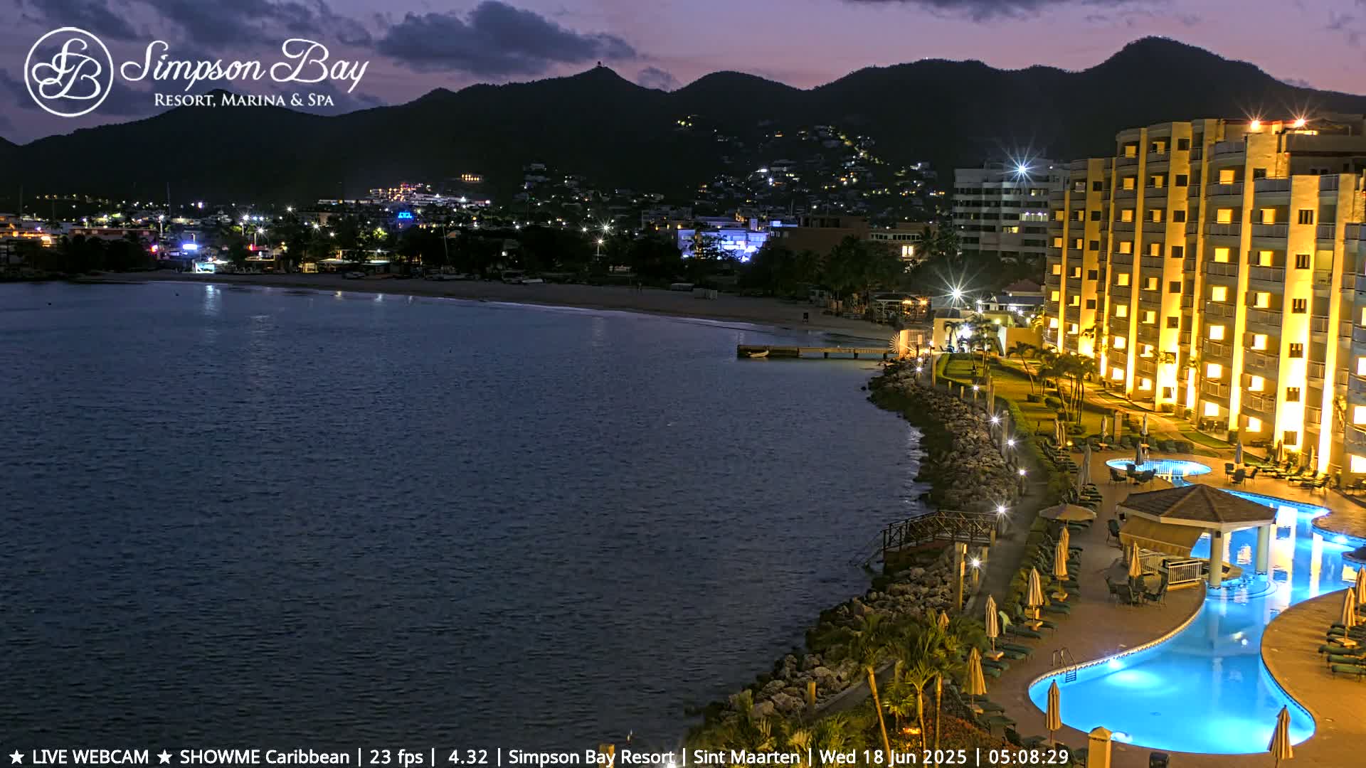 A brightly lit resort with a large, curved pool sits on a rocky shoreline at twilight, overlooking a calm bay and a dark mountain range in the distance.