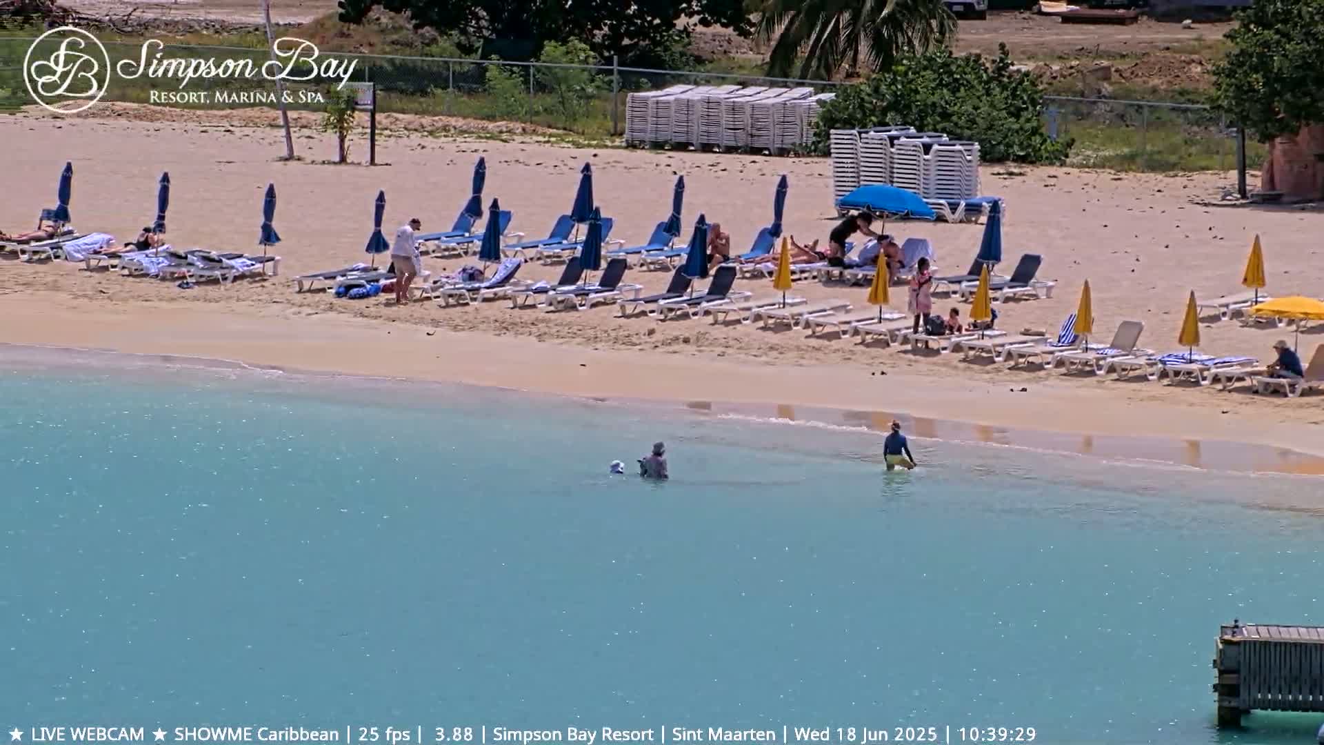 A sandy beach with lounge chairs and umbrellas, some people sunbathing and swimming in calm, turquoise water.