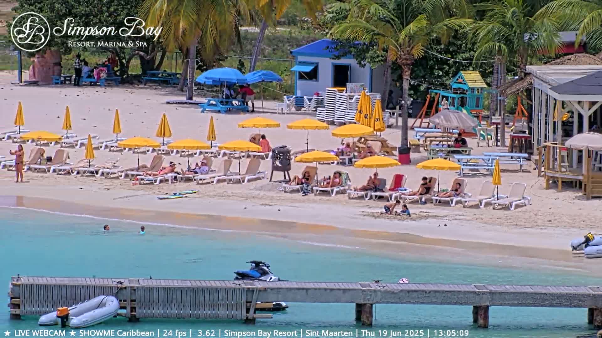A sunny beach scene shows numerous yellow umbrellas and lounge chairs on a sandy beach, with turquoise water, a dock, and a few people swimming and relaxing.