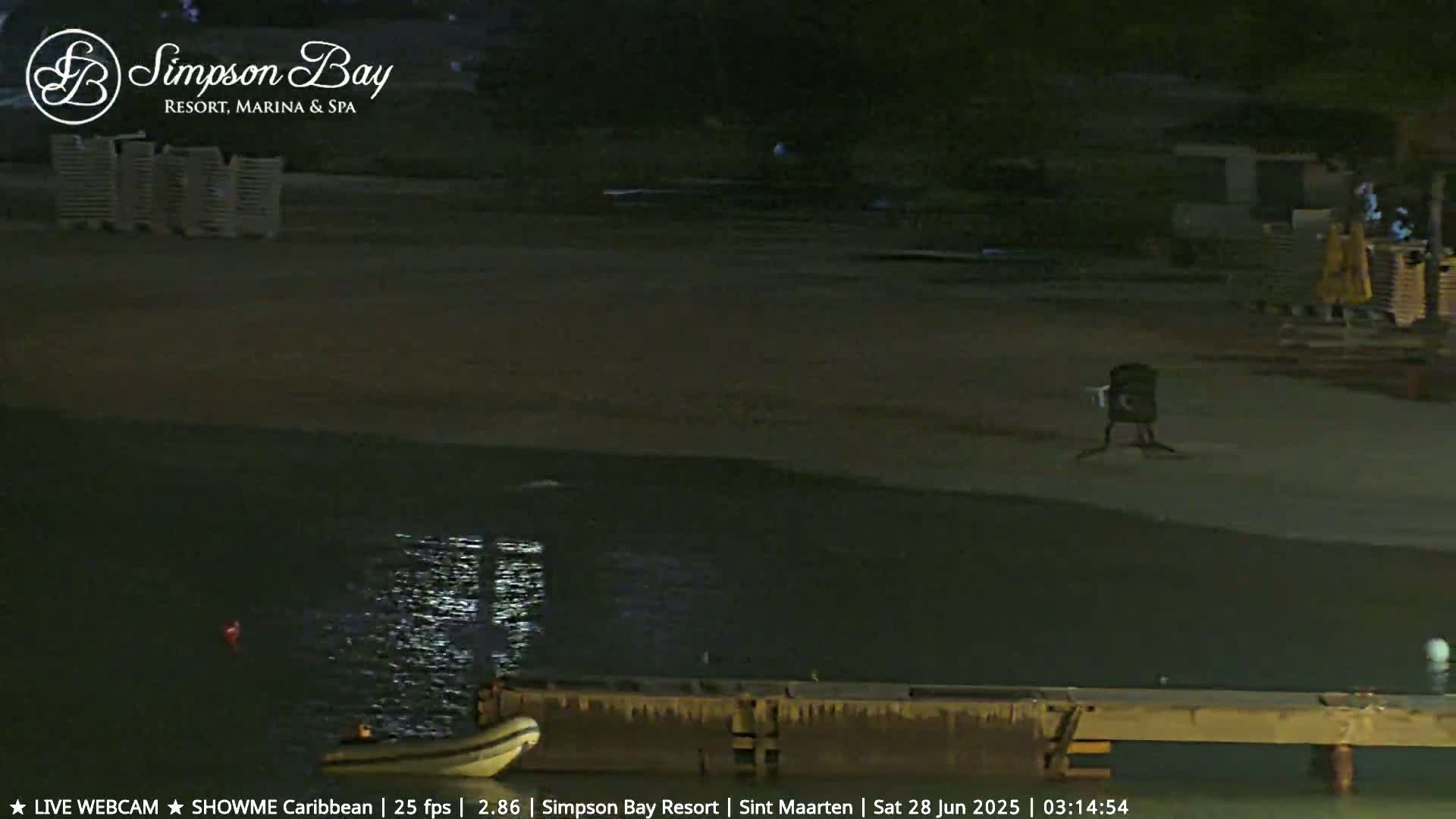 A dark, nighttime beach scene shows a calm body of water next to a mostly empty sandy shore with a small boat at a dock.
