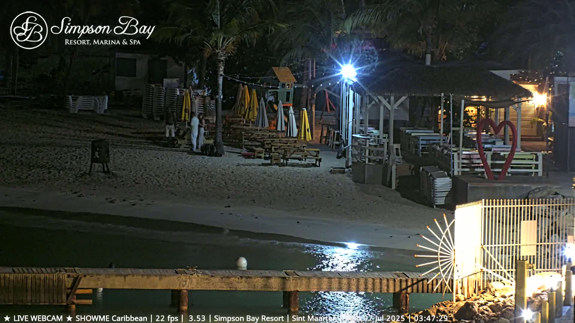 A nighttime beach scene shows a sandy shore with a beachfront restaurant under a thatched roof, several picnic tables, and umbrellas; calm water laps gently at the shore.