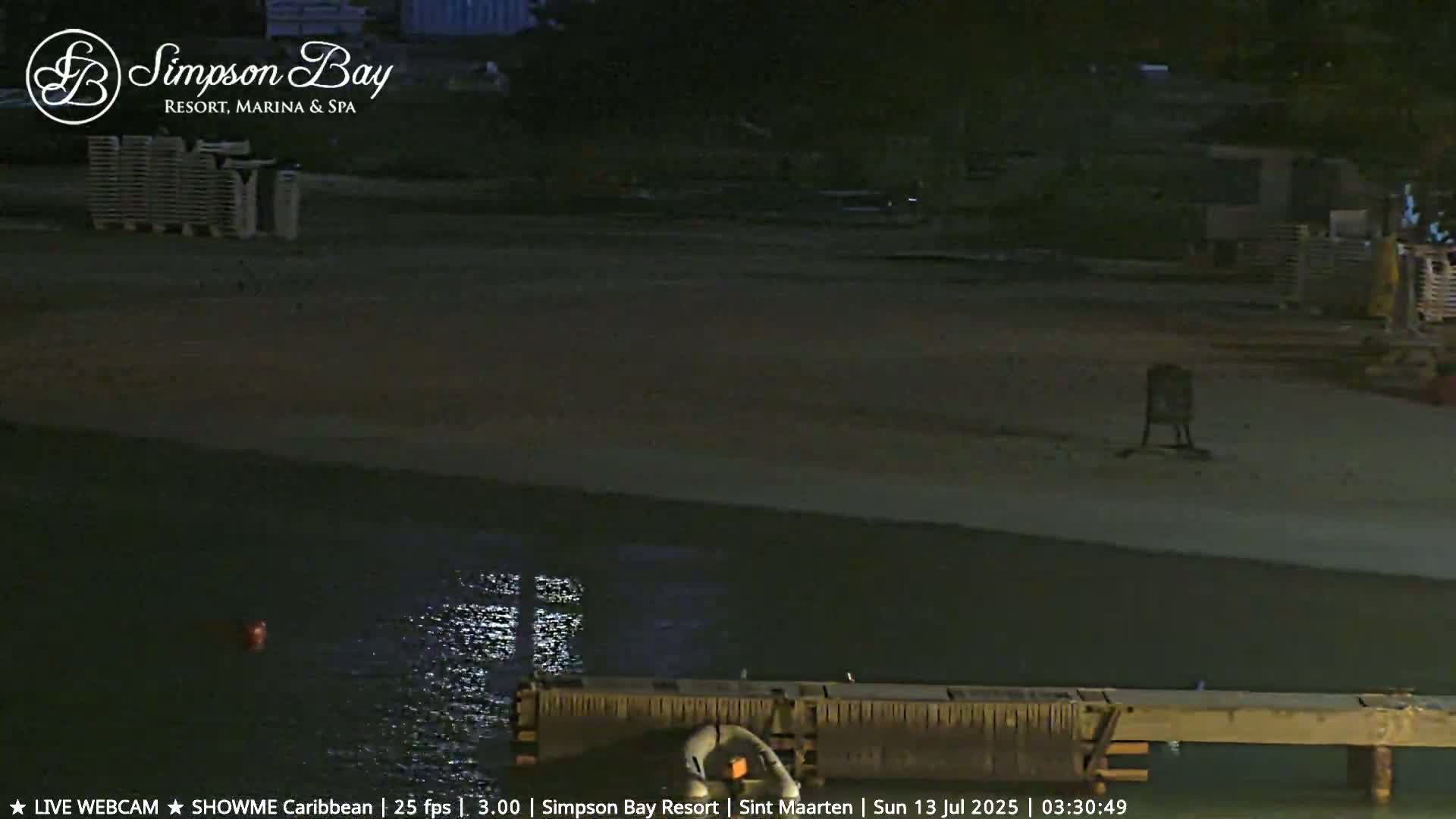 A dark beach at night is shown with calm water, a dock, and stacked lounge chairs in the background.