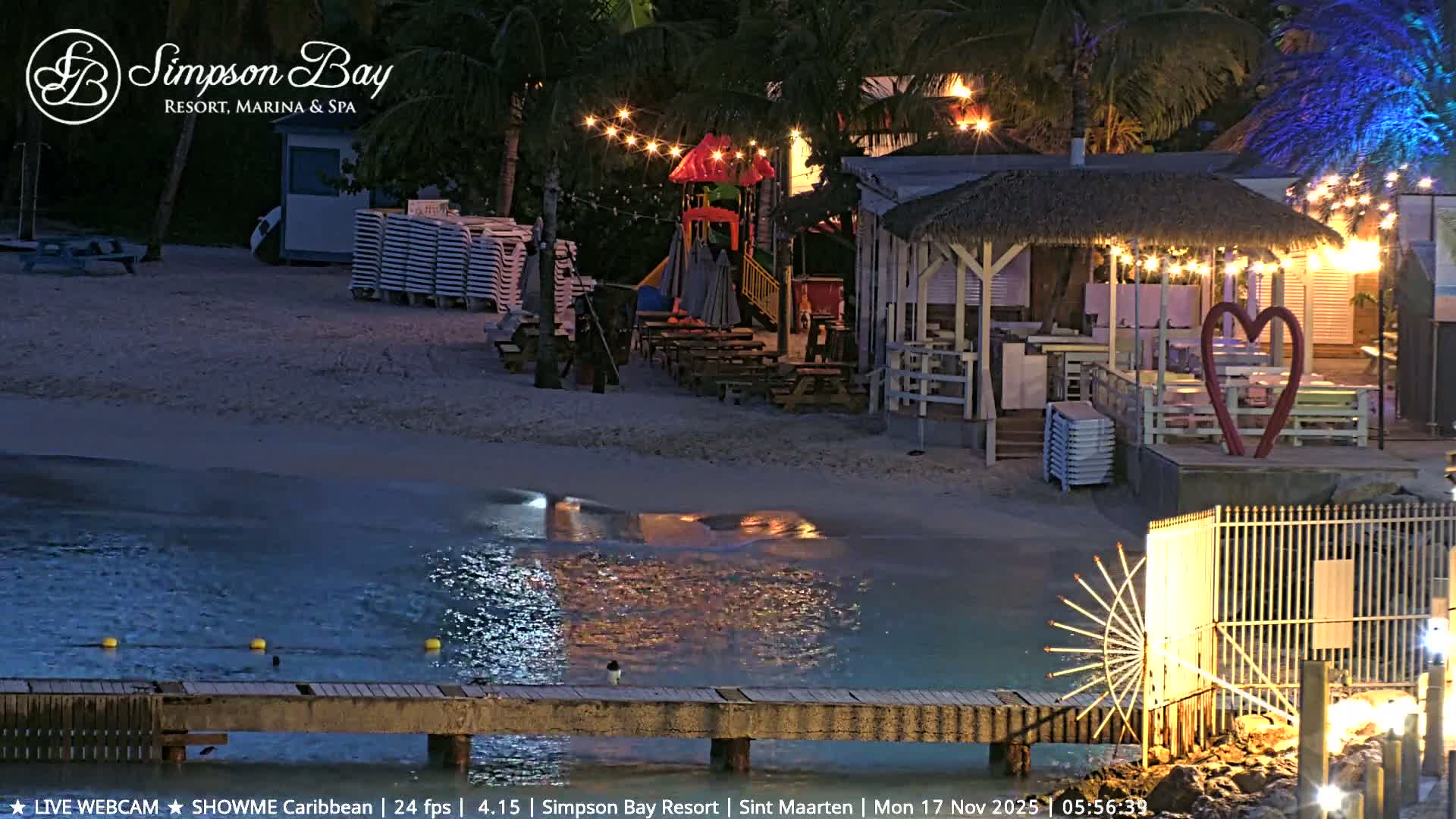 A tranquil resort beach is depicted at night under clear skies, featuring a wooden pier extending into calm waters reflecting string lights, alongside palm trees, a colorful children's playground, a thatched-roof bar, stacked loungers, and a large red heart sculpture on the sandy shore.