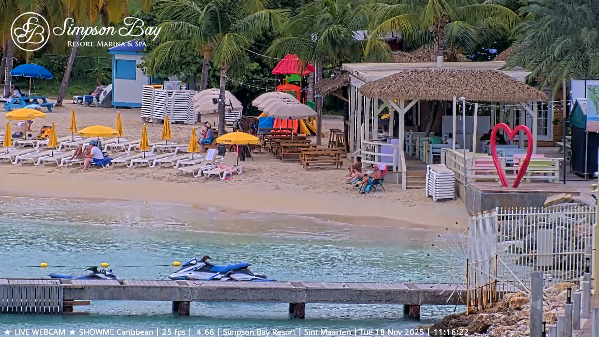 A tranquil resort beach is depicted at night under clear skies, featuring a wooden pier extending into calm waters reflecting string lights, alongside palm trees, a colorful children's playground, a thatched-roof bar, stacked loungers, and a large red heart sculpture on the sandy shore.