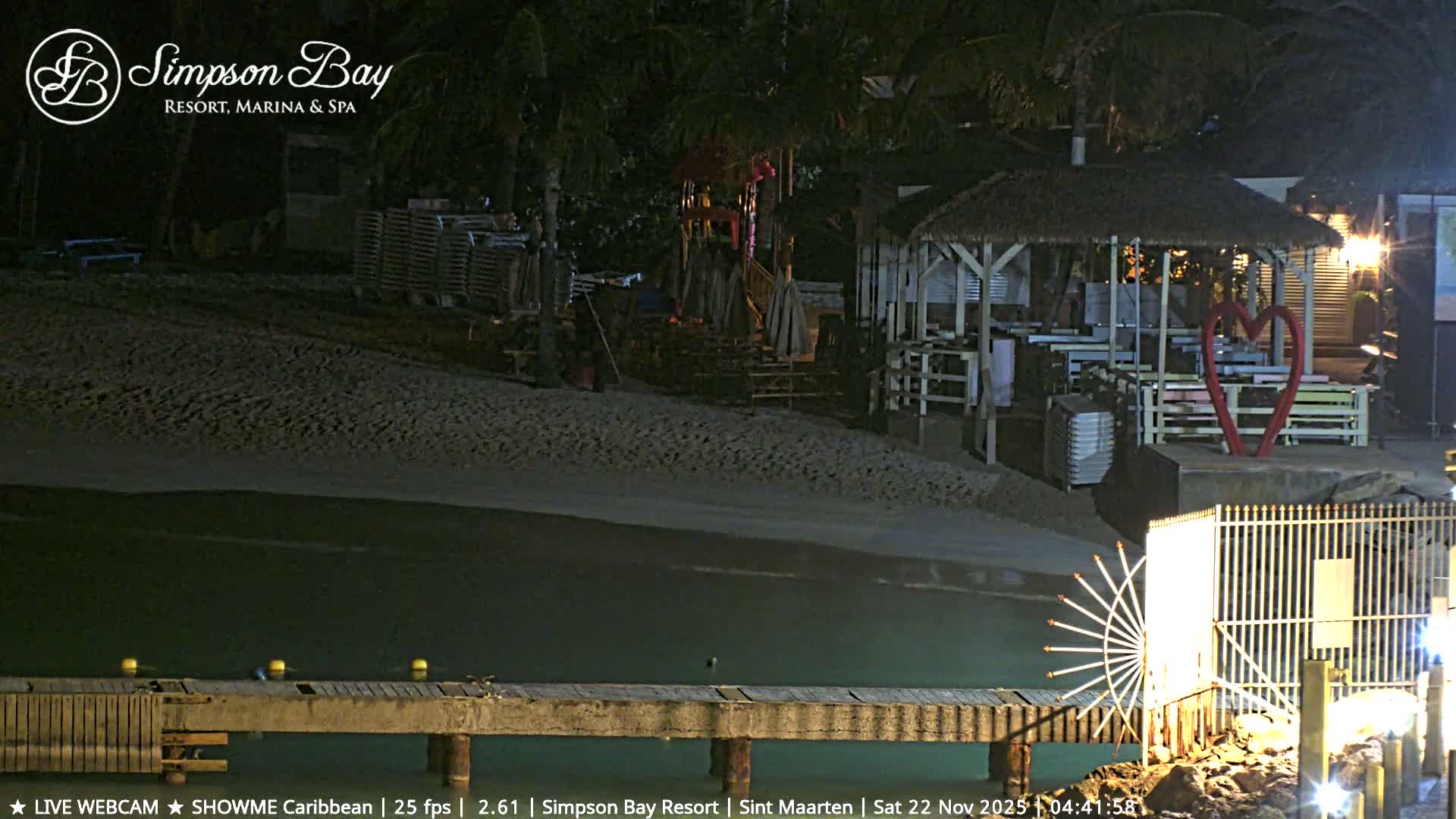 This nighttime image depicts a tranquil resort beach scene with a wooden pier extending into calm water, a sandy shore, and various illuminated structures including a thatched-roof hut, dining areas, and a large red heart sculpture, all under clear skies.