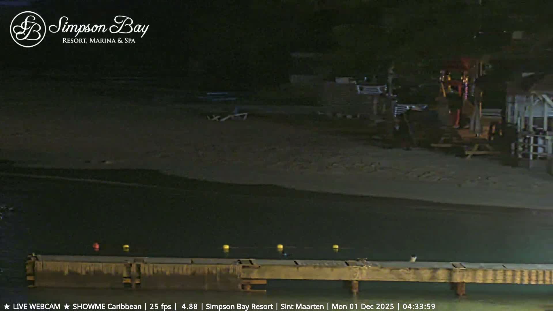 The image displays a clear, dark night at a beach with a wooden pier extending into the water where several colorful buoys float, and a sandy shore featuring dimly lit beach structures, picnic tables, and lounge chairs.