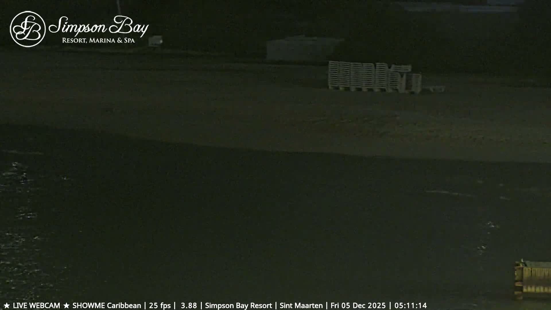 The dimly lit image shows a tranquil outdoor scene at night, featuring calm water in the foreground, a sandy shore with stacked pallets, and a faint structure in the distance under clear skies.