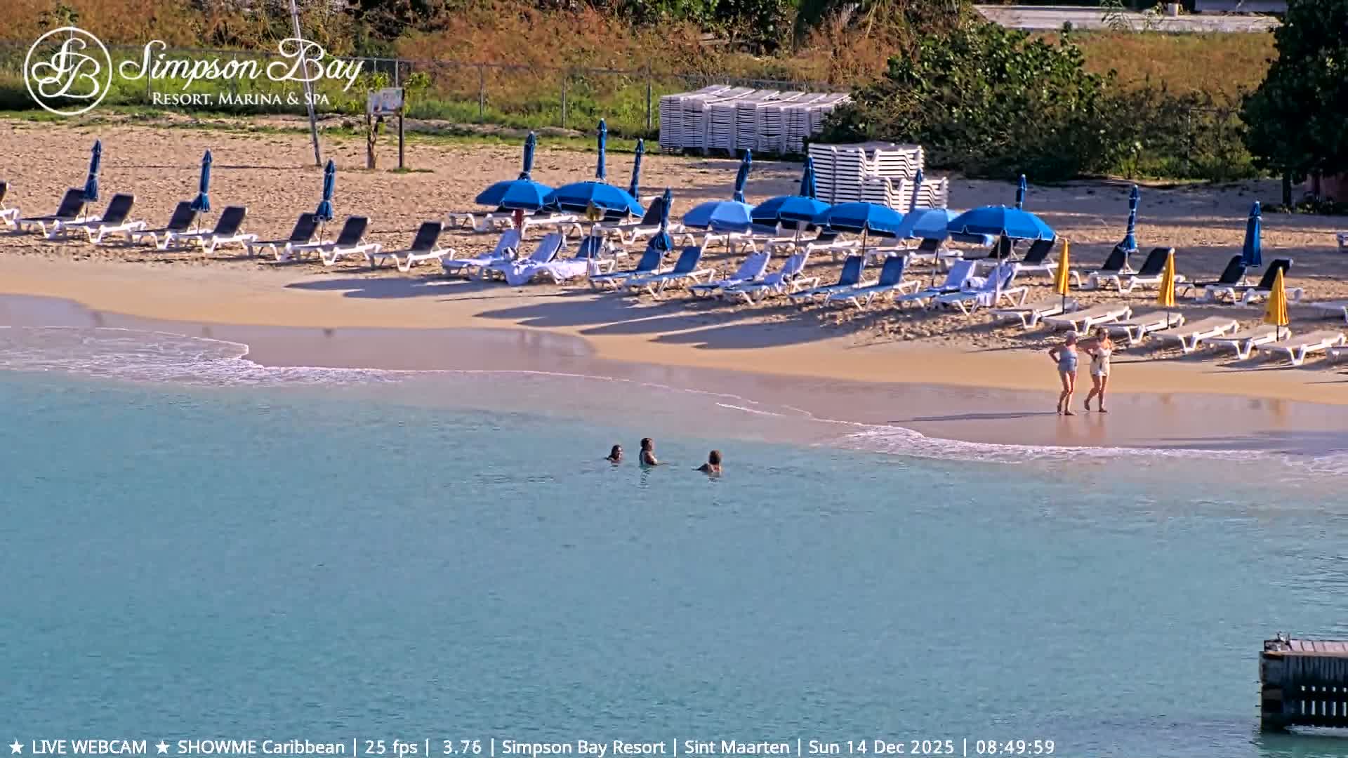 The dimly lit image shows a tranquil outdoor scene at night, featuring calm water in the foreground, a sandy shore with stacked pallets, and a faint structure in the distance under clear skies.