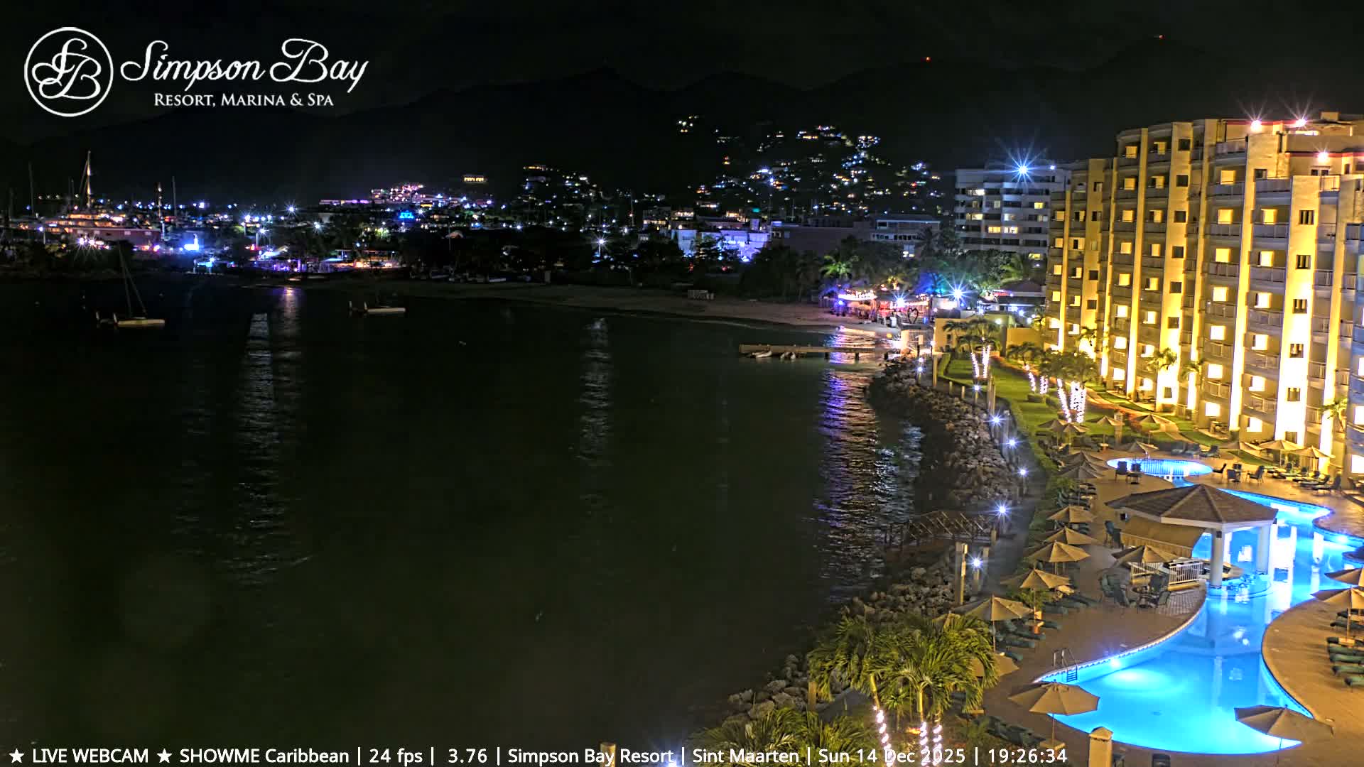 The dimly lit image shows a tranquil outdoor scene at night, featuring calm water in the foreground, a sandy shore with stacked pallets, and a faint structure in the distance under clear skies.