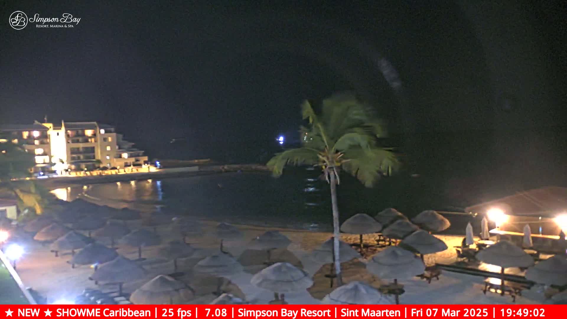 A nighttime beach scene shows a resort building, palm trees, and numerous closed beach umbrellas under a clear, dark sky.