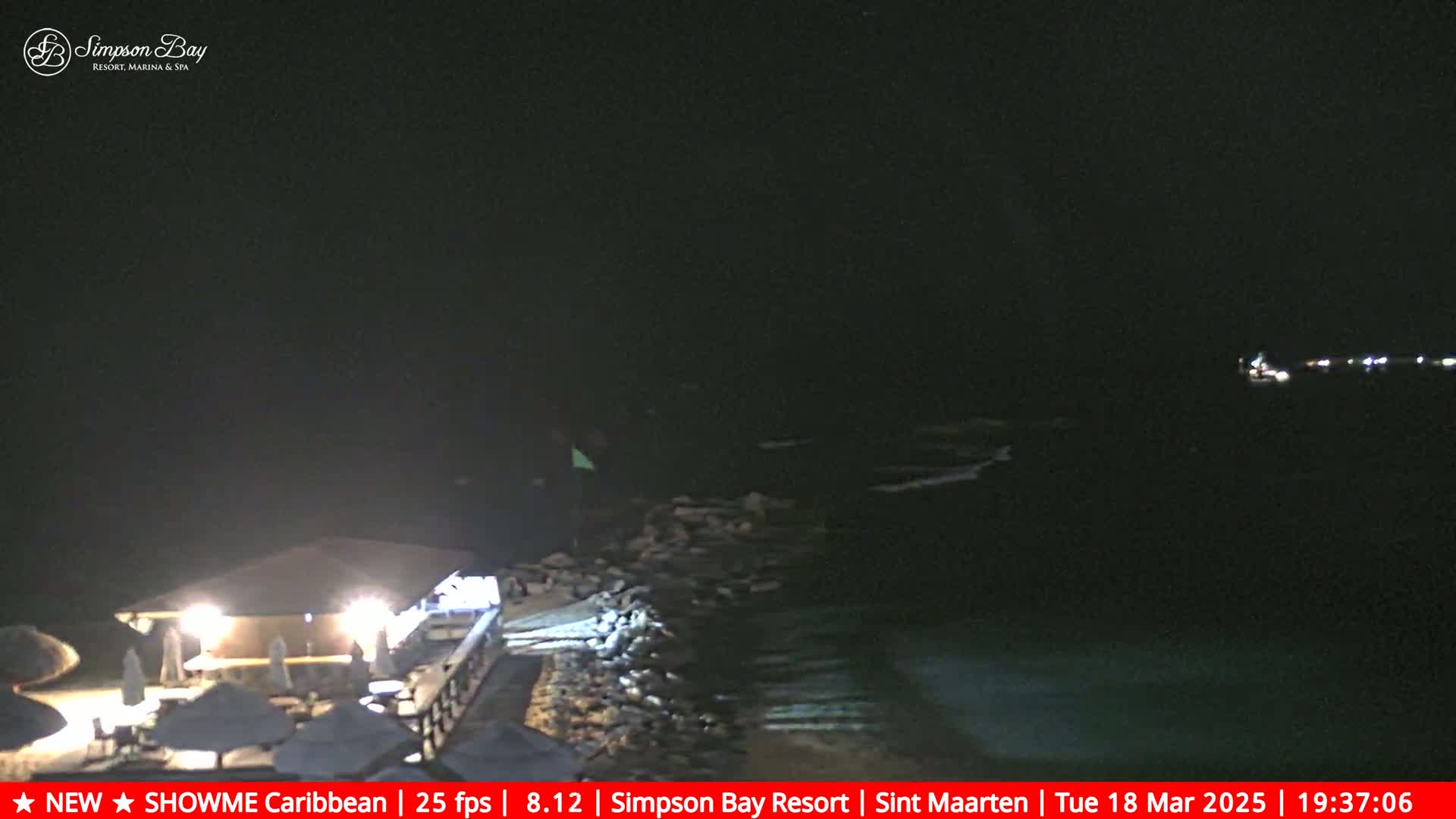 A nighttime view of a dark ocean with a lit-up beachfront restaurant and some small boats visible in the distance.