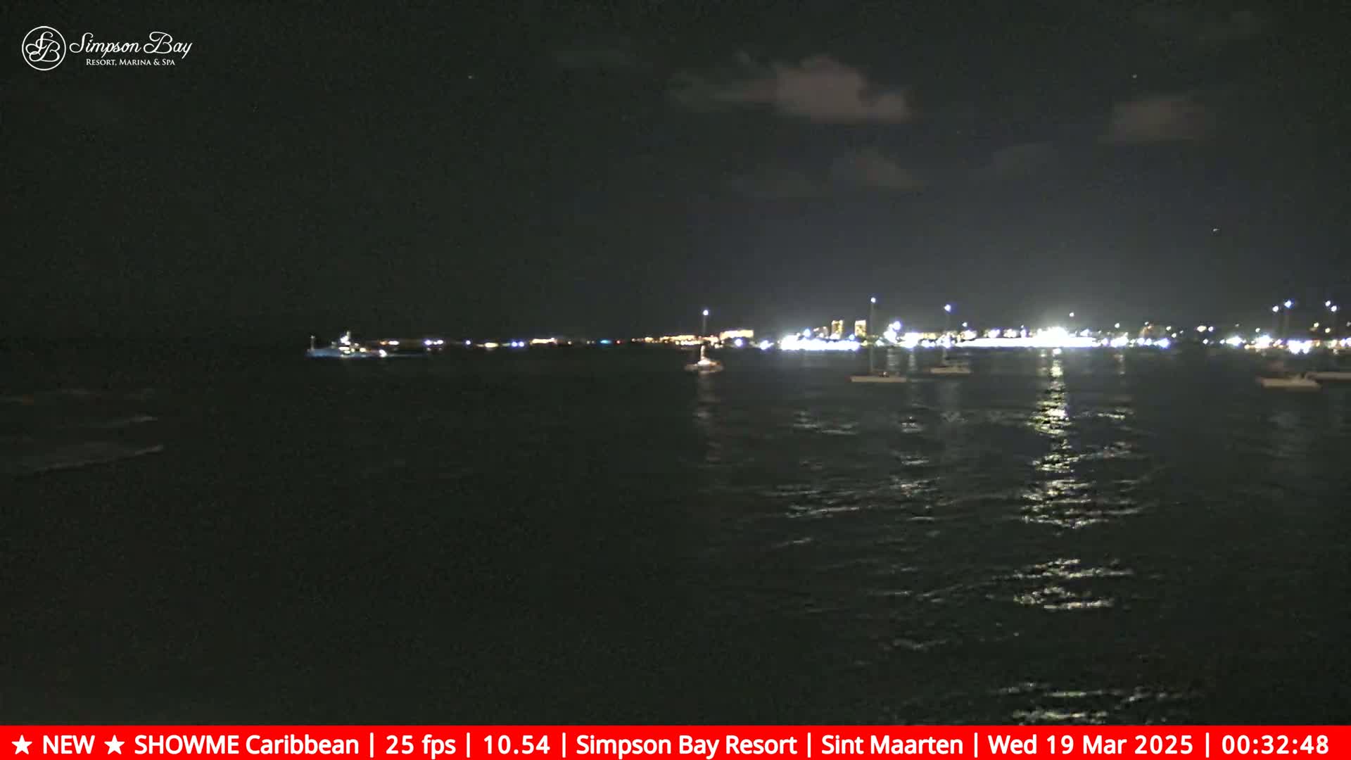 A nighttime view of a calm body of water with several boats and a distant, illuminated shoreline under a partly cloudy sky.