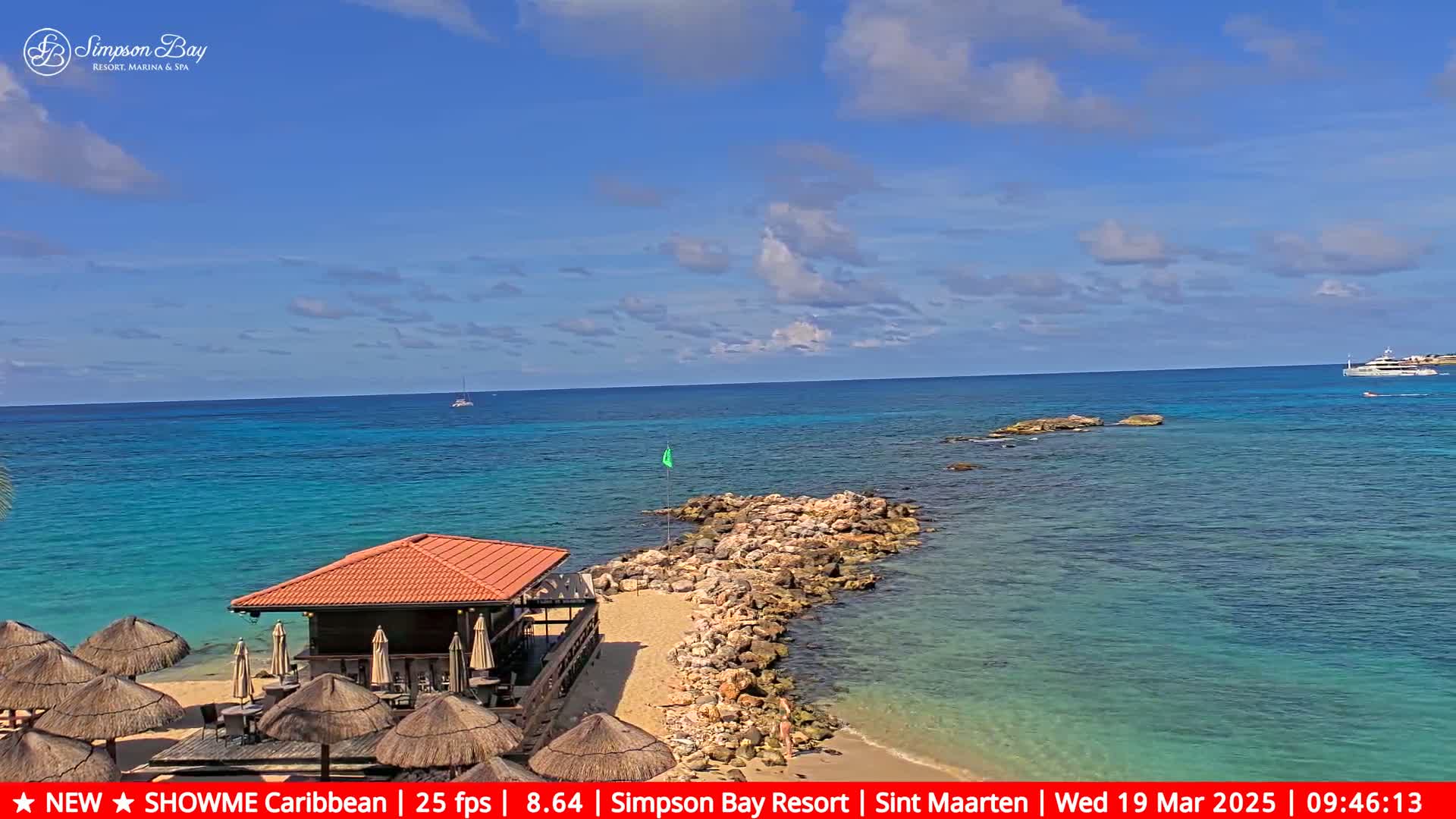 A beach bar with thatched-roof umbrellas sits on a sandy beach next to a rocky jetty extending into a calm, turquoise ocean under a mostly sunny sky.