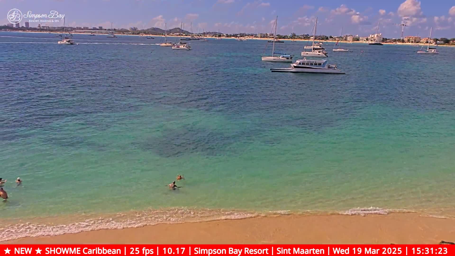 Several people swim in clear, shallow turquoise water near a sandy beach, with sailboats and other vessels visible in the calm ocean beyond.