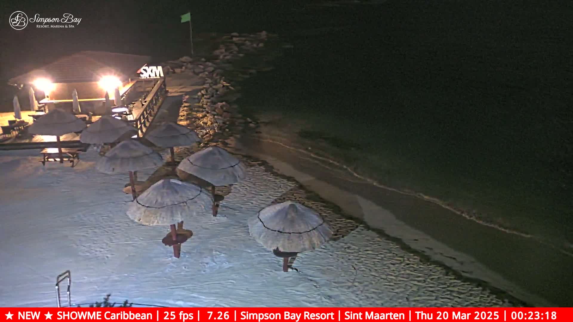 A nighttime beach scene shows several thatched umbrellas on a sandy shore near a lit beachfront bar, with gentle waves lapping the sand under a dark sky.