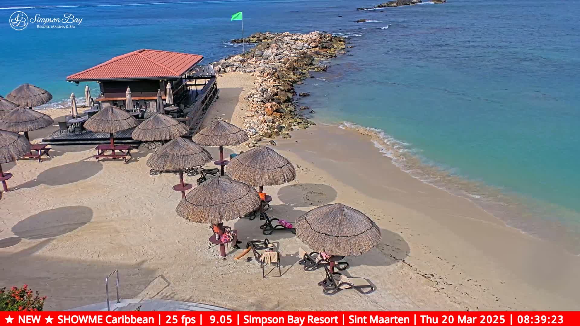 A sandy beach with thatched umbrellas and beach chairs sits next to a small bar, all bordered by a rocky outcropping and a calm, turquoise ocean under a sunny sky.