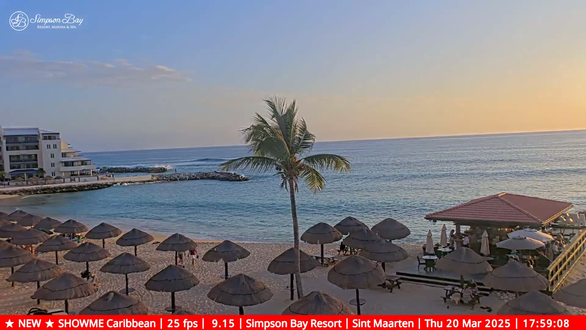 A sandy beach with numerous thatched umbrellas, a beachfront bar, and calm ocean under a clear, sunset sky.