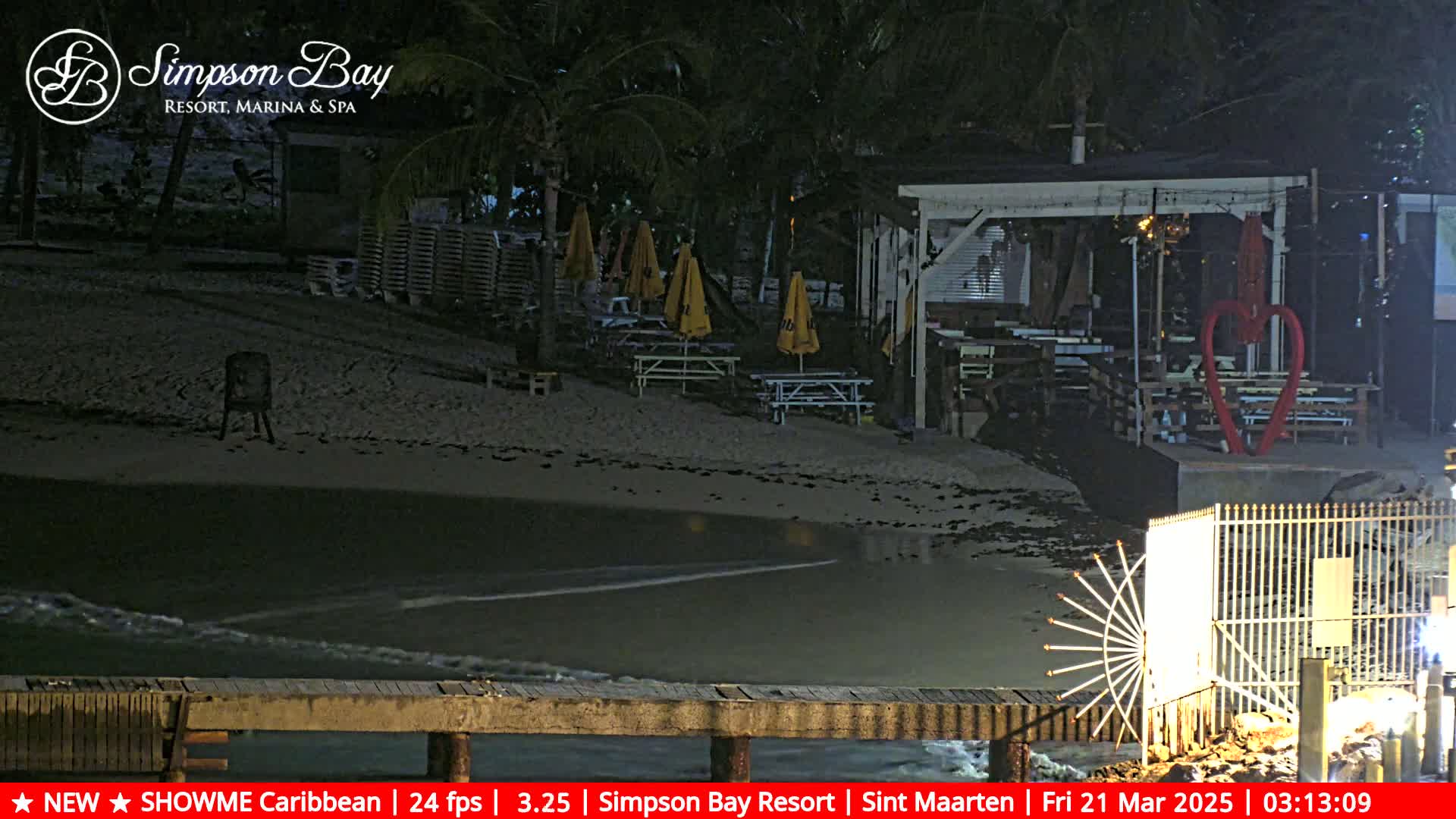 A nighttime beach scene shows a calm body of water lapping at a sandy shore, with beach furniture and a building under dim lighting.