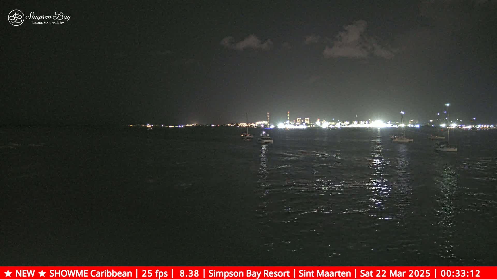 A nighttime view of a dark body of water with several boats and a brightly lit cityscape in the distance under a partly cloudy sky.