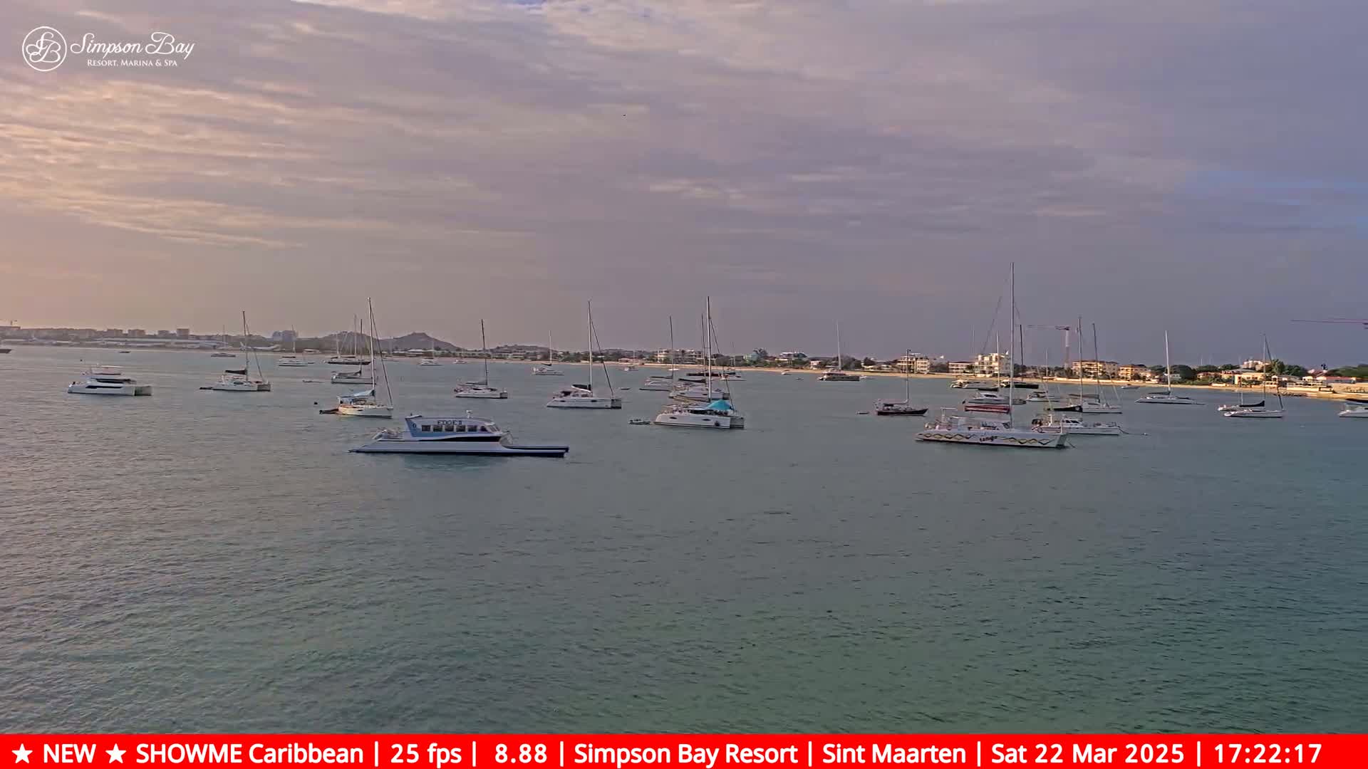 Many boats are anchored in calm water near a shoreline with buildings under a partly cloudy sky.