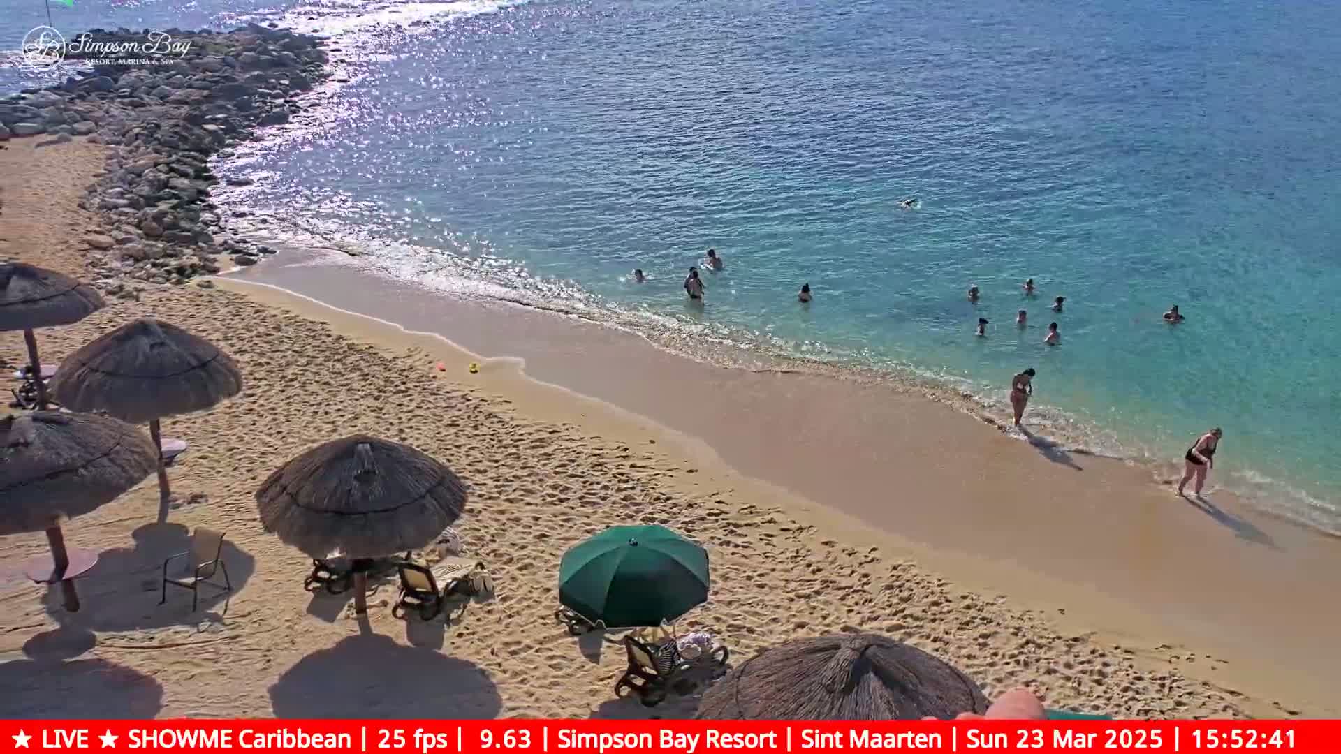 A sandy beach with several thatched umbrellas and a green umbrella, a calm, clear blue ocean with people swimming, and a rocky shoreline.