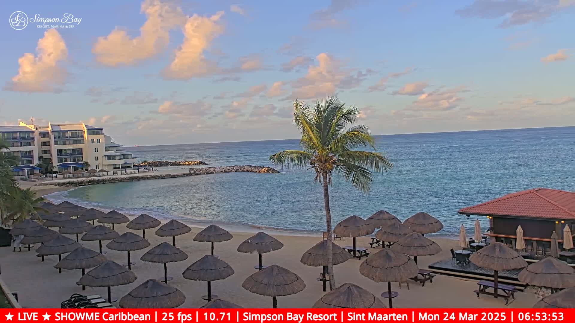 A sandy beach with numerous thatched umbrellas and a beachfront bar is visible under a partly cloudy sky, next to calm ocean water and a resort building.