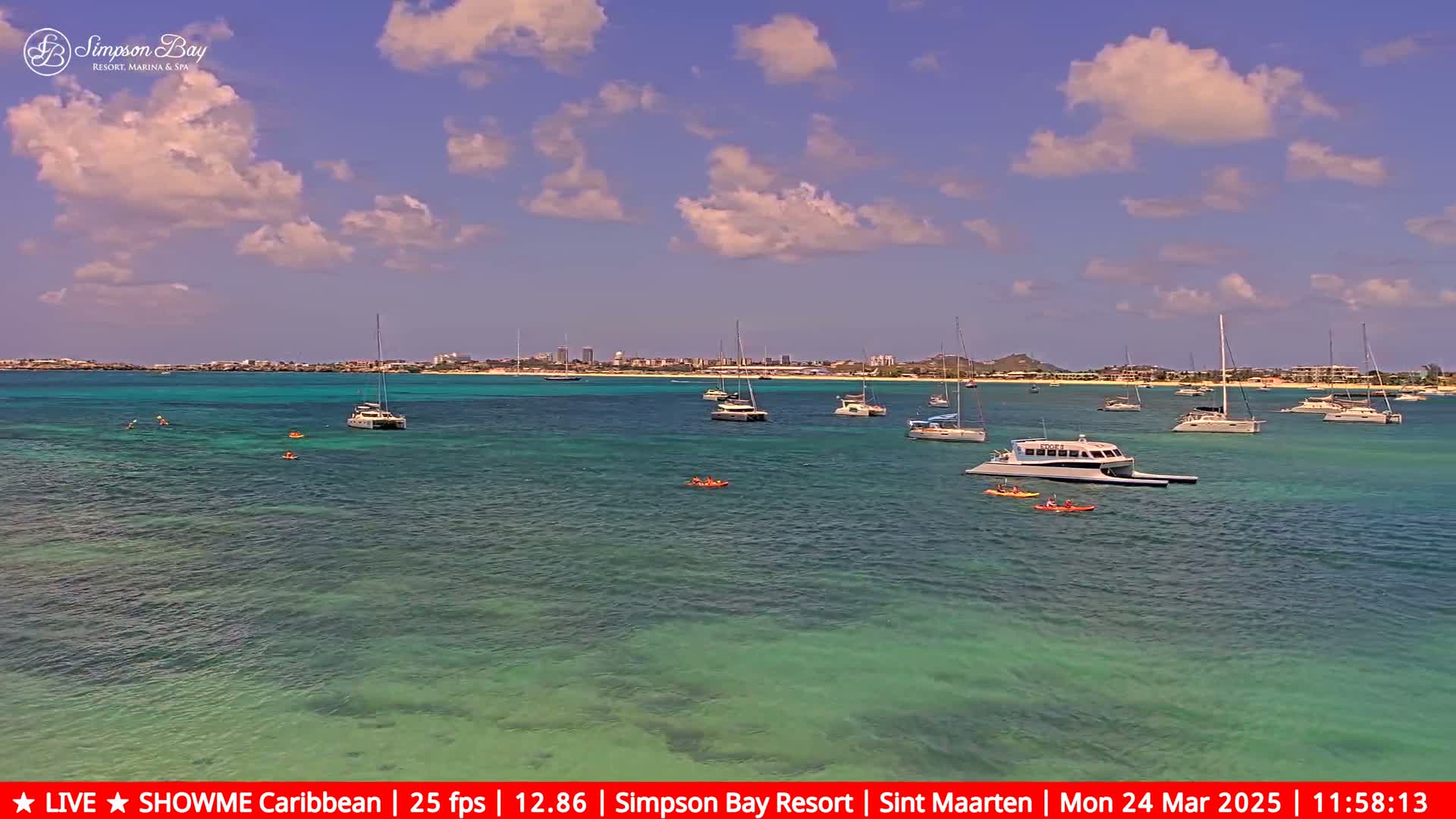 Several sailboats and motorboats are anchored in clear, shallow turquoise water under a partly cloudy sky.