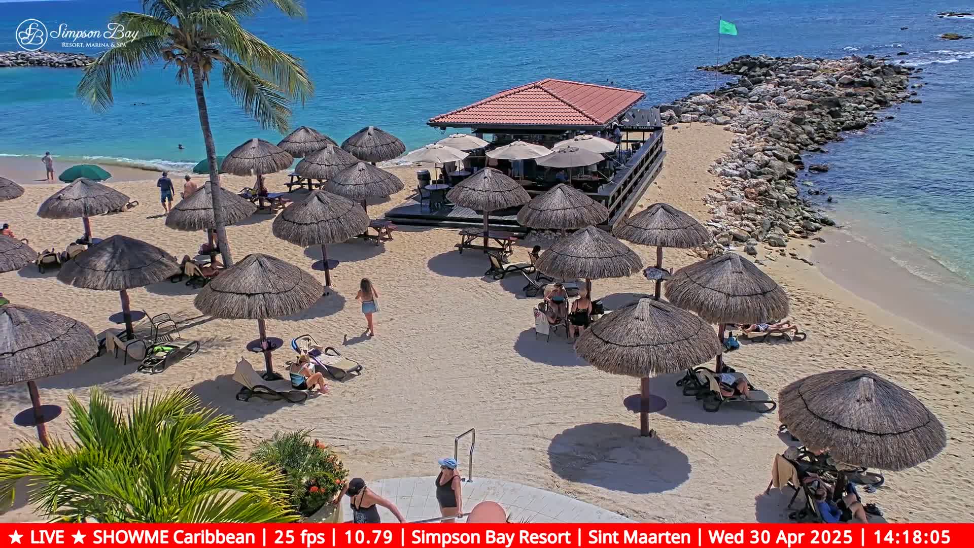 A sandy beach with numerous thatched umbrellas, lounge chairs, and people relaxing under a sunny sky next to a small beach bar and calm, clear ocean.